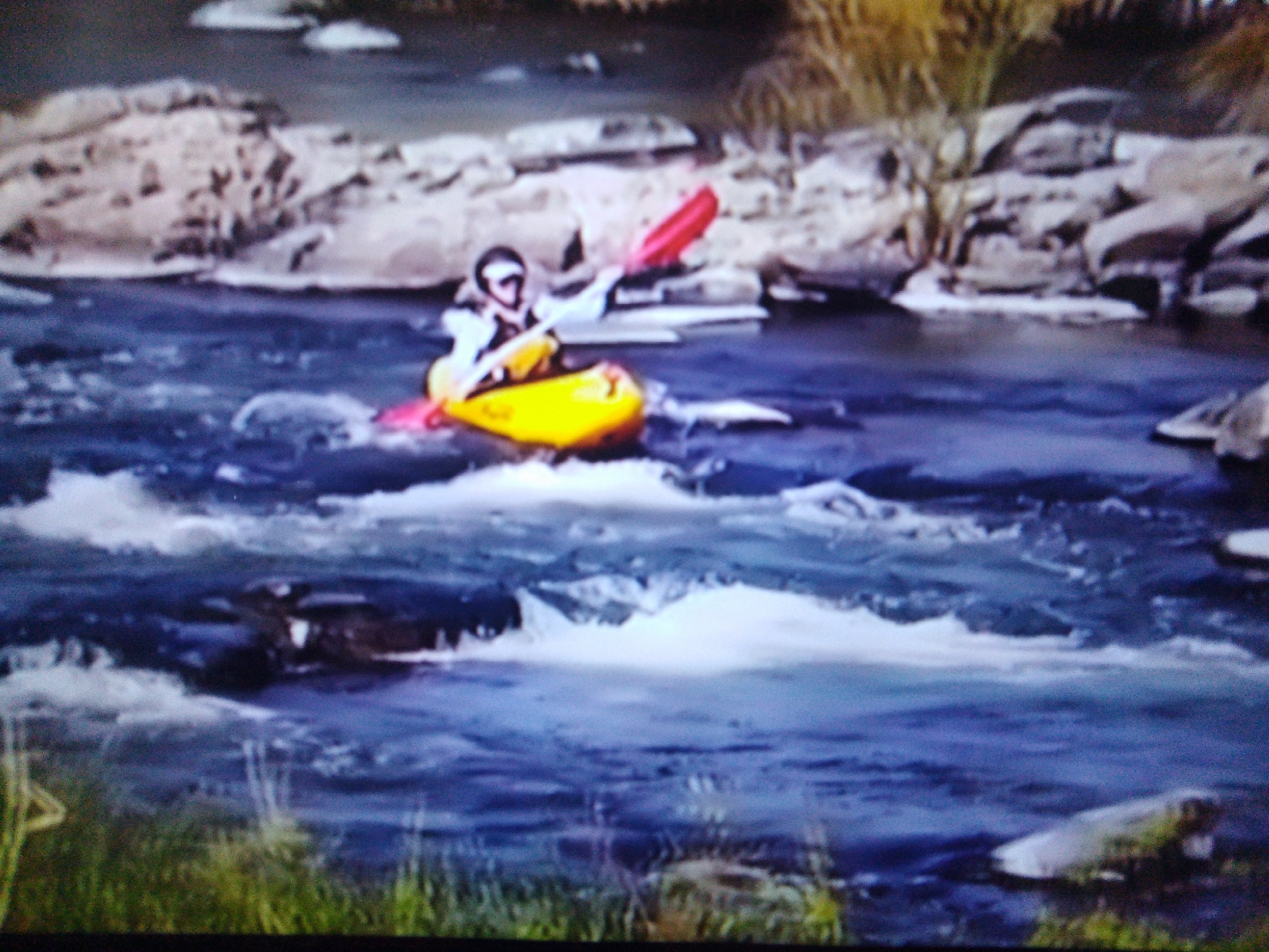 A person wearing a helmet and life vest is kayaking down a turbulent river. The kayaker is in a yellow kayak and holding a red paddle. The river has rocky banks and vegetation. The scene appears to be a wilderness setting. The image is blurry, suggesting it may be a photograph taken from a distance or a low-quality video frame. There is no visible indication of the city of Dushanbe, Tajikistan.
