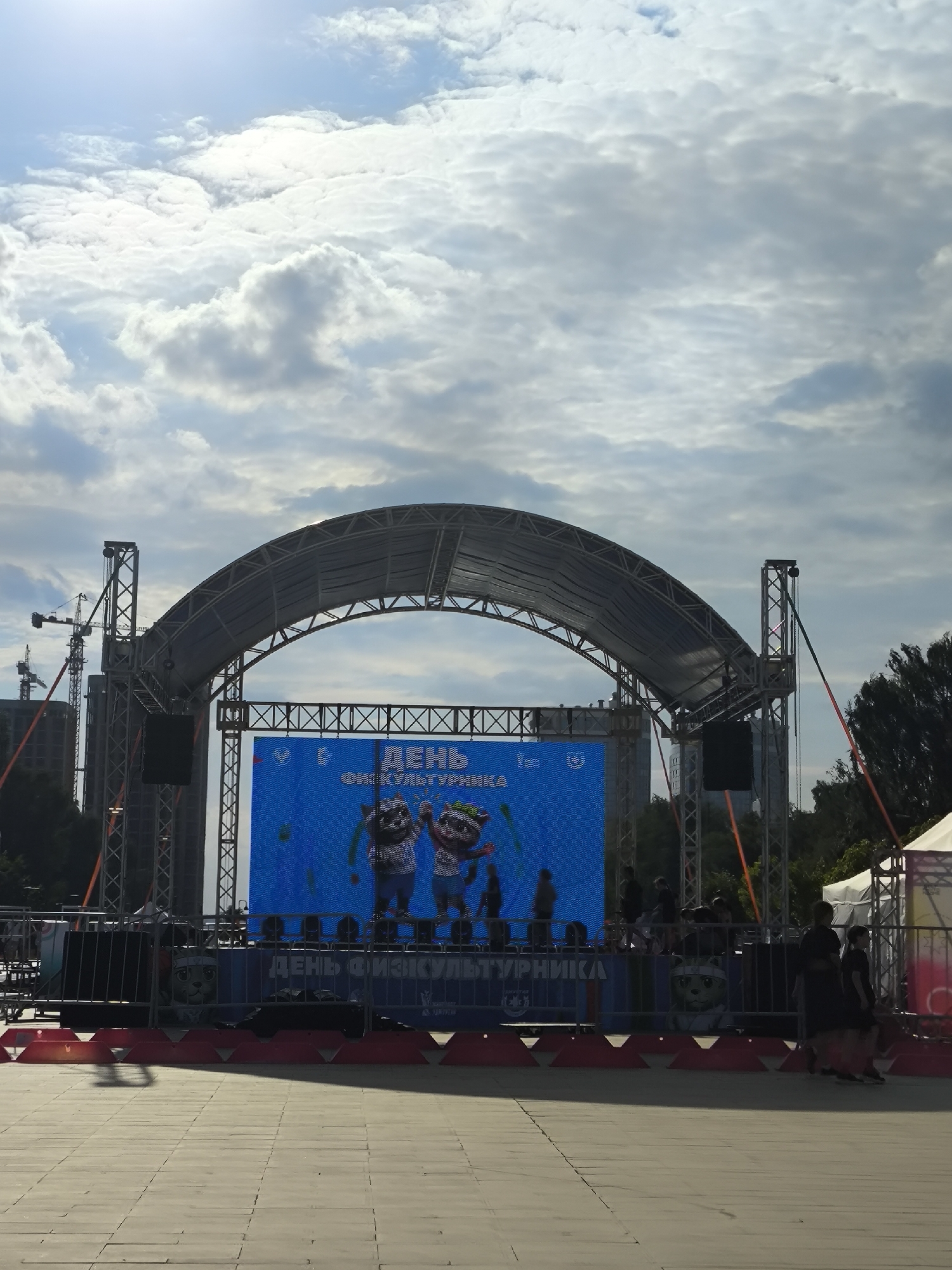 An outdoor stage with a large LED screen is set up in a paved public space. The screen displays a blue background with cartoon characters and text that appears to be in Cyrillic. A curved metal structure covers the stage. In the background, construction cranes and tall buildings are visible. Several people are present, some standing near the stage and others walking in the foreground. The sky is filled with clouds. This scene is located on the central square of Izhevsk, Russia, in front of the "Udmurtia" government building, during a public event.