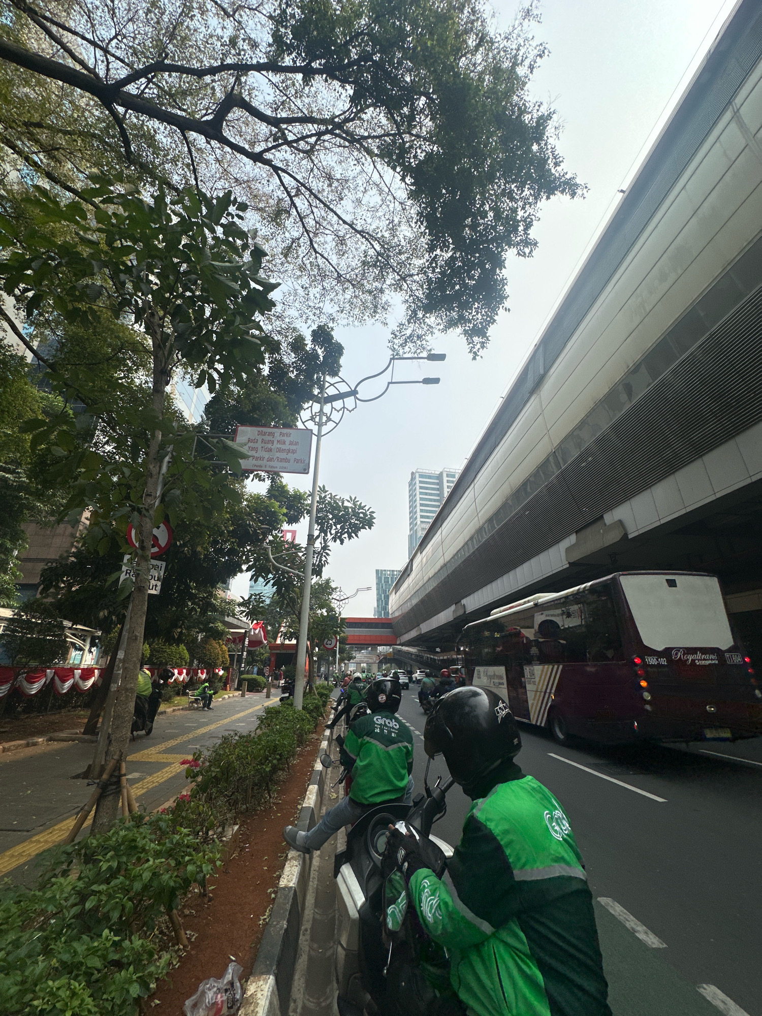 The image depicts a street scene in South Jakarta, Indonesia, likely on a bright, possibly hazy day. In the foreground, two individuals wearing green uniforms with "Grab" branding are on motorcycles. One is closer to the camera, facing away, with a black helmet. The other is slightly further back, also on a motorcycle. They appear to be waiting or stopped.
To the left, a sidewalk with red and white decorative banners lines the street. Trees with green foliage are present, and a traffic sign indicating "Dilarang Parkir" (No Parking) is visible on a pole, along with another circular red and white traffic sign. Further down the street, other vehicles, including motorcycles and cars, are visible.
Dominating the background is a large, elevated structure, likely an overpass or a section of an elevated train or highway. A maroon bus with "Royaltrans" branding is traveling on the road beneath this structure. In the distance, modern city buildings rise above the trees. The overall impression is of a busy urban environment with transportation infrastructure.