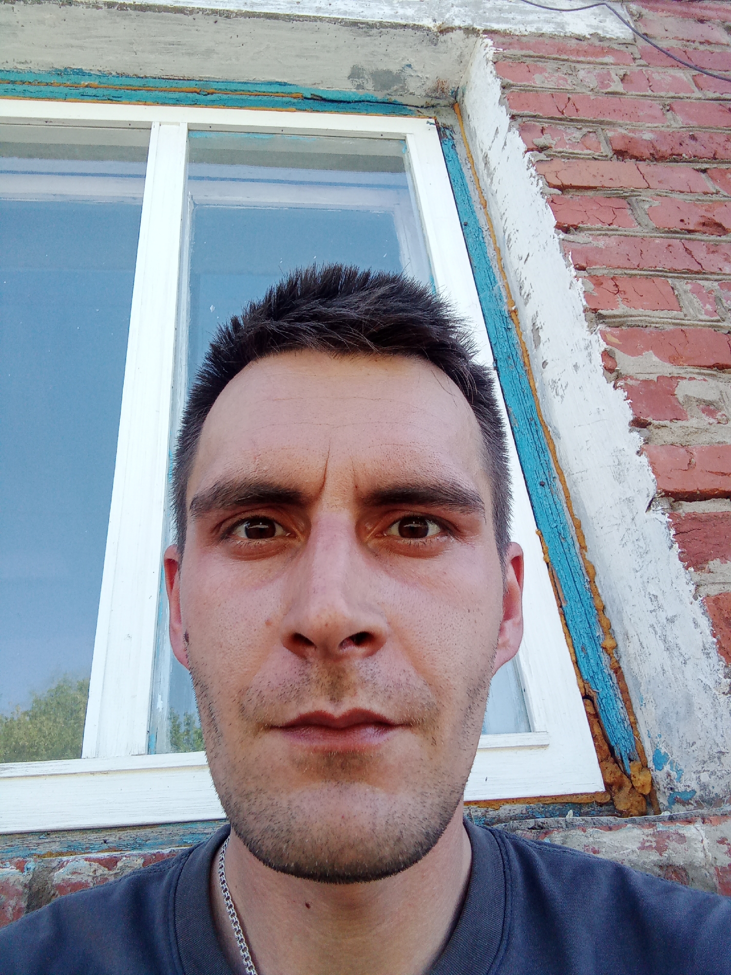 A man with short, dark hair is centered in the frame, facing the camera. He has brown eyes, a medium complexion, and stubble. He wears a dark-colored T-shirt and a silver chain is visible around his neck. He appears to be frowning slightly.
Behind him is a window with a white frame, reflecting the sky. To the right of the window, a red brick wall is visible with peeling white paint. Blue paint is also present on the window frame and surrounding areas.
While it is impossible to definitively confirm the location in the city of Chaikovsky, Russia based solely on the image, the presence of a worn, older window frame, a brick wall, and a certain style of construction could be consistent with the architectural styles found in some areas of Russia. However, without further information or identifying features, it's impossible to confirm.