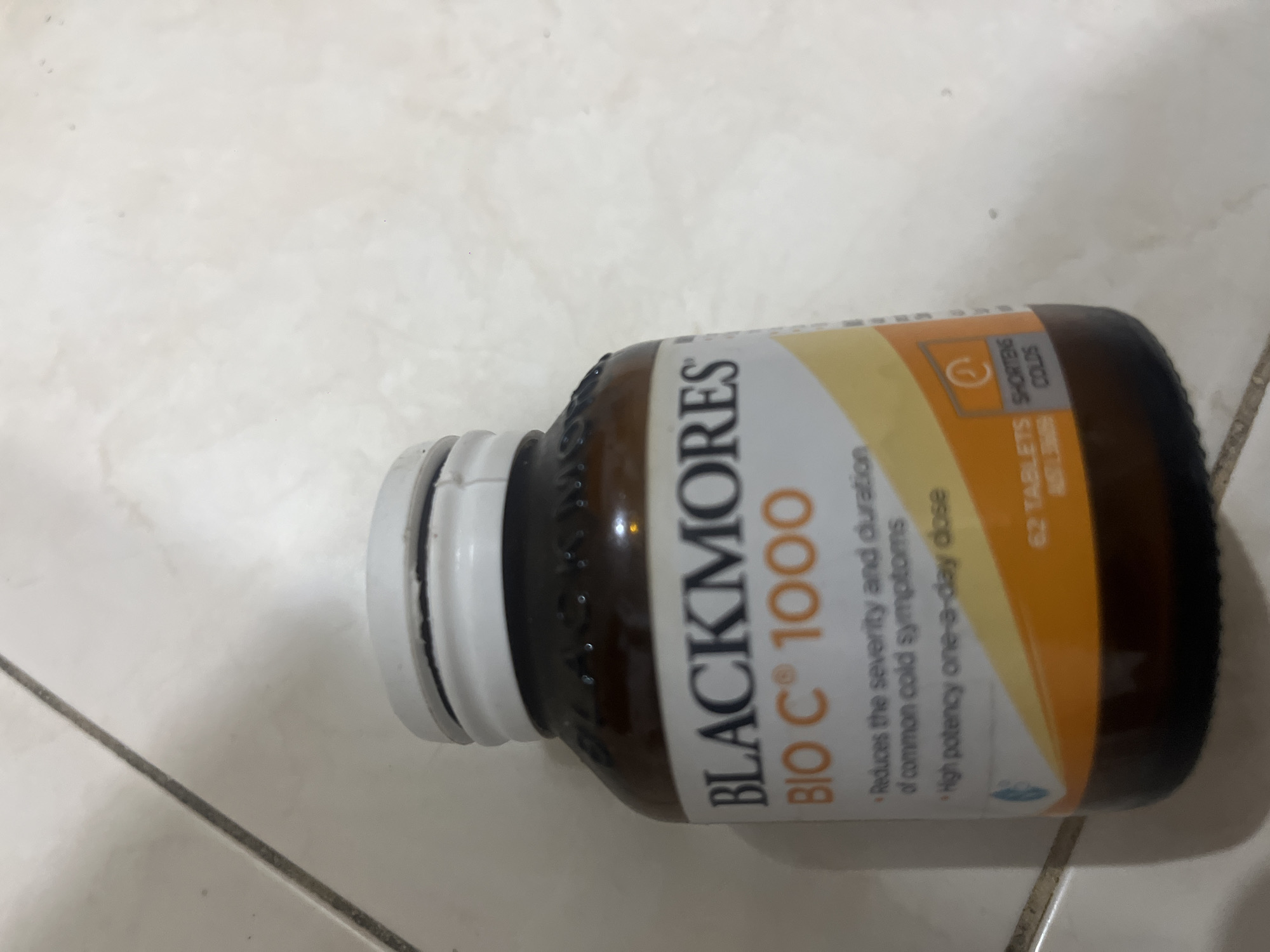 The image shows a close-up of a bottle of Blackmores Bio C 1000 tablets, placed on a tiled surface. The bottle is amber glass with a white plastic lid. The label on the bottle is white and orange, with text indicating the product name, "BIO C® 1000," and claims such as "Reduces the severity and duration of common cold symptoms" and "High potency one-a-day dose." The bottom of the label has text that includes "62 TABLETS" and "SHORTENS COLDS." The background is a tiled floor with visible grout lines. There is no indication of the city of Denpasar, Indonesia, in the image.