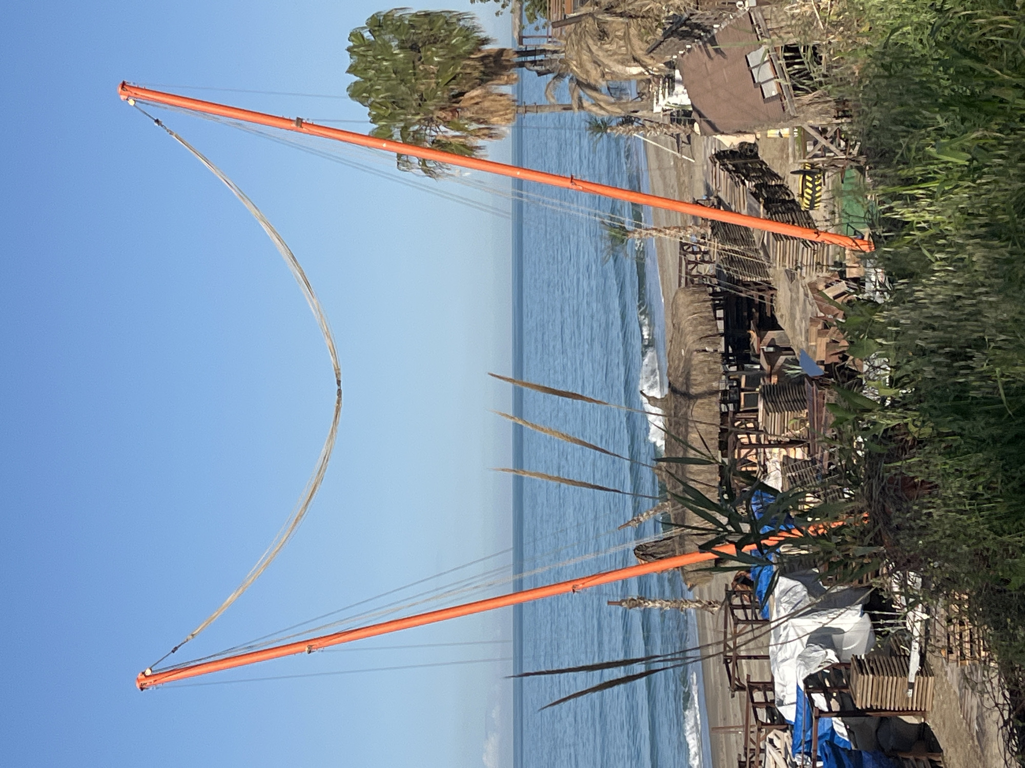 The image depicts a beach scene in Alanya, Turkey, featuring a bungee jumping apparatus. Two orange metal arms extend upwards from a structure on the beach, connected by cables that form a large arc. The beach is visible with the ocean in the background under a clear blue sky. The shoreline is marked by small waves. Debris, including chairs, building materials and what appears to be covered furniture, are scattered along the beach near the base of the apparatus. Trees and vegetation are also present. The bungee apparatus is positioned near a building, possibly a beachfront establishment. The overall scene suggests a public beach area with recreational facilities. No people are visible in the image.