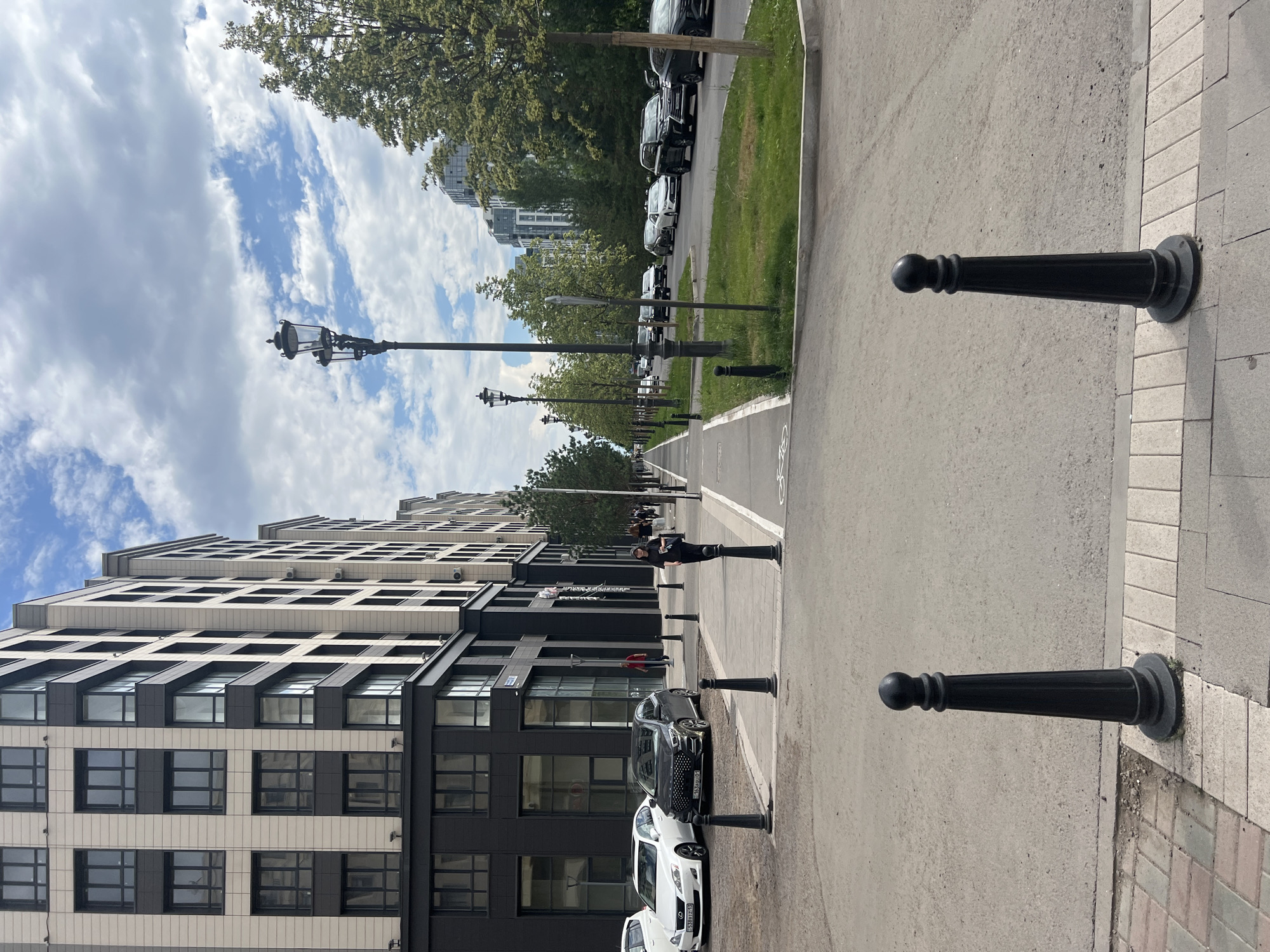 The image captures a pedestrian walkway and a street in Astana, Kazakhstan, during daylight. Modern, multi-story buildings with predominantly glass windows and light-colored facades line the left side of the street. Several cars are parked along the street and in traffic. A person in dark clothing is walking on the pedestrian path. Streetlights with a classic design are positioned along the walkway. Trees with green foliage are planted on the right side of the street, and grass verges separate the road from the pedestrian area. The sky is partly cloudy with a mix of blue sky and white clouds. The ground is paved with asphalt and cobblestones.