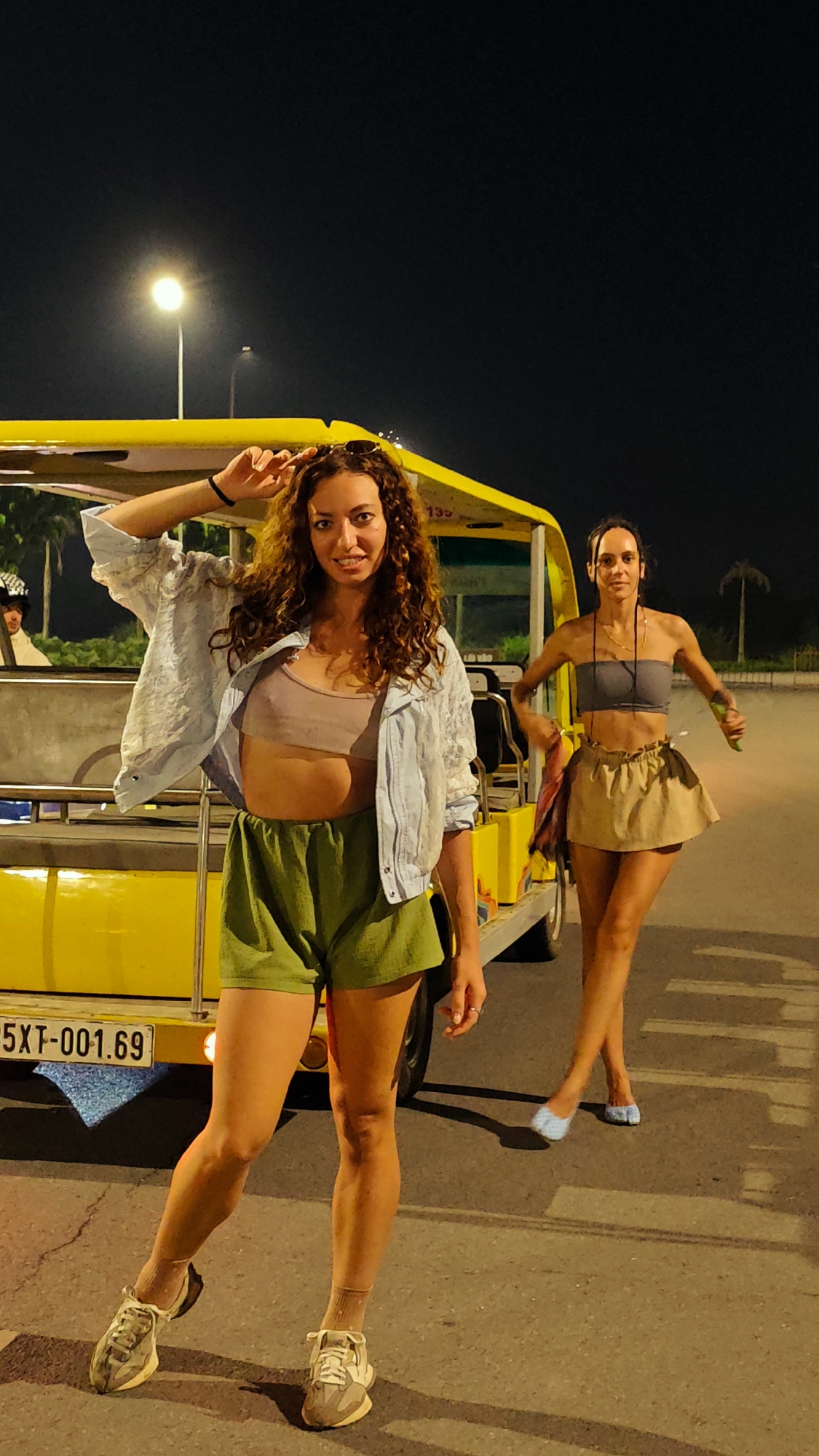 Two women stand in front of a yellow vehicle at night in Tây Hoa Lư Ward, Vietnam. The woman in the foreground, on the left, has long, curly brown hair and is wearing a light blue denim jacket over a light purple crop top and green shorts. She is also wearing white sneakers and beige socks. Her left hand is raised to her head. The woman in the background, on the right, has dark hair pulled back and is wearing a grey strapless top and a tan skirt. She is wearing blue slip-on shoes. The yellow vehicle appears to be a small bus or tram. A license plate on the vehicle reads "5XT-001.69". The background is dark with a streetlamp visible to the left and palm trees to the right, suggesting an outdoor, possibly tourist area.