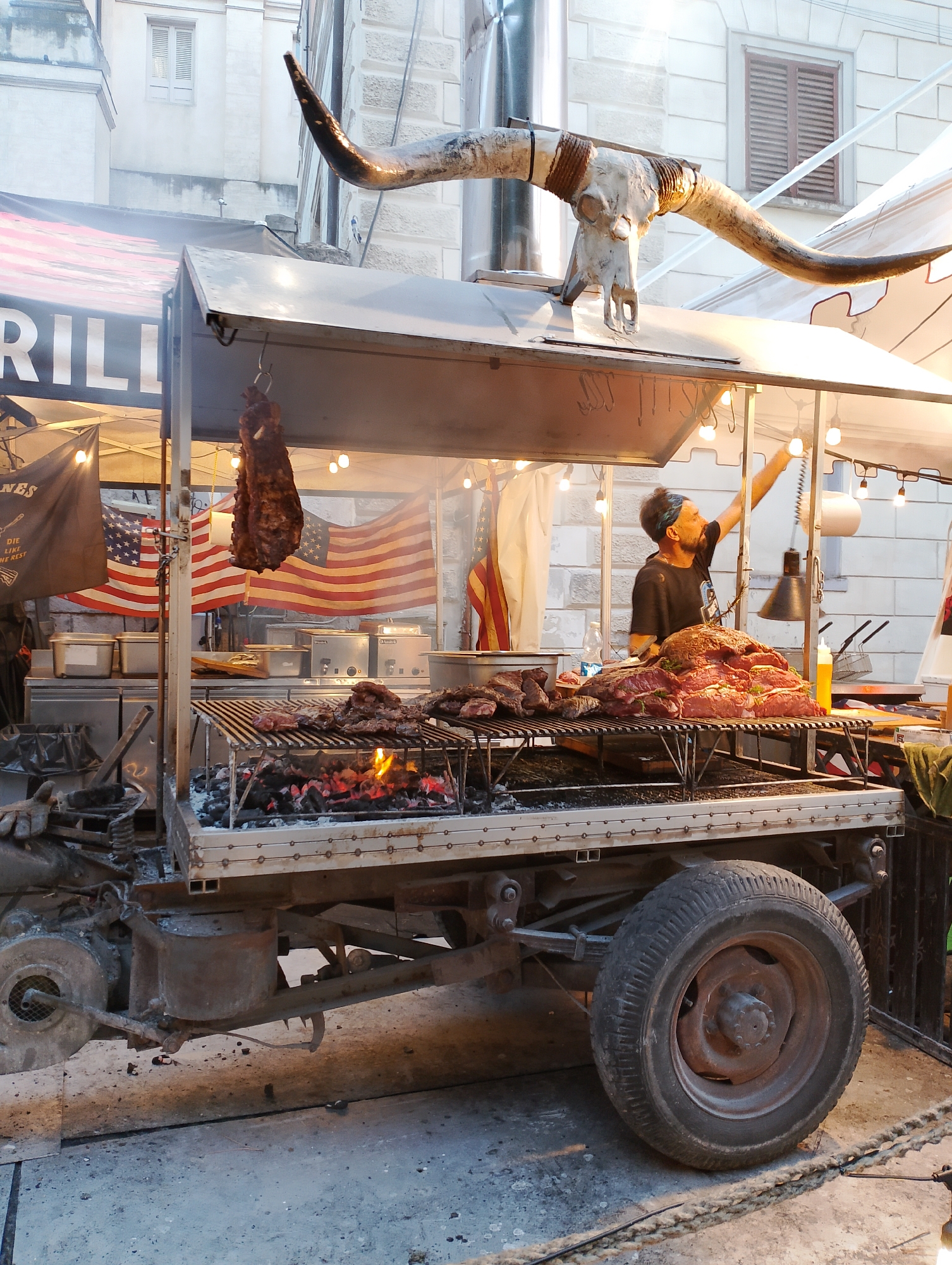 The image captures a vibrant scene at a street food vendor, possibly in Rieti, Italy, given the provided context. Dominating the foreground is a large, mobile grill setup, likely a food truck or cart, adorned with a striking bull skull and longhorns as decoration. The grill itself is active, with glowing coals and several cuts of meat cooking over the heat. A man, presumably the vendor, is visible in the background, tending to the grill and interacting with equipment. American flags are prominently displayed behind the grill, adding a thematic element to the setup. The environment suggests an outdoor setting, possibly a market or a street fair, with buildings and lights visible in the background. The overall impression is one of a lively, gastronomic establishment.