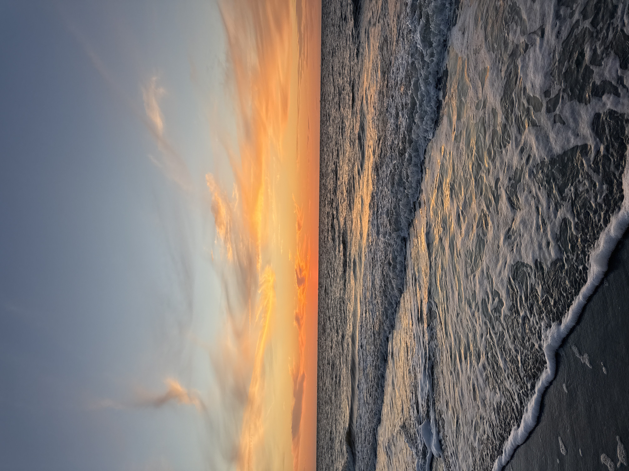 The image depicts a serene ocean sunset at the beach on Langeoog, Germany. The sky is a gradient of soft blues and vibrant oranges, with wispy clouds catching the warm light. The sun is low on the horizon, casting a golden glow across the water. Gentle waves are rolling onto the sandy shore, creating a frothy white foam along the edge of the water. The overall scene is peaceful and tranquil, characteristic of a sunset at the beach. There are no people or specific objects in the frame.