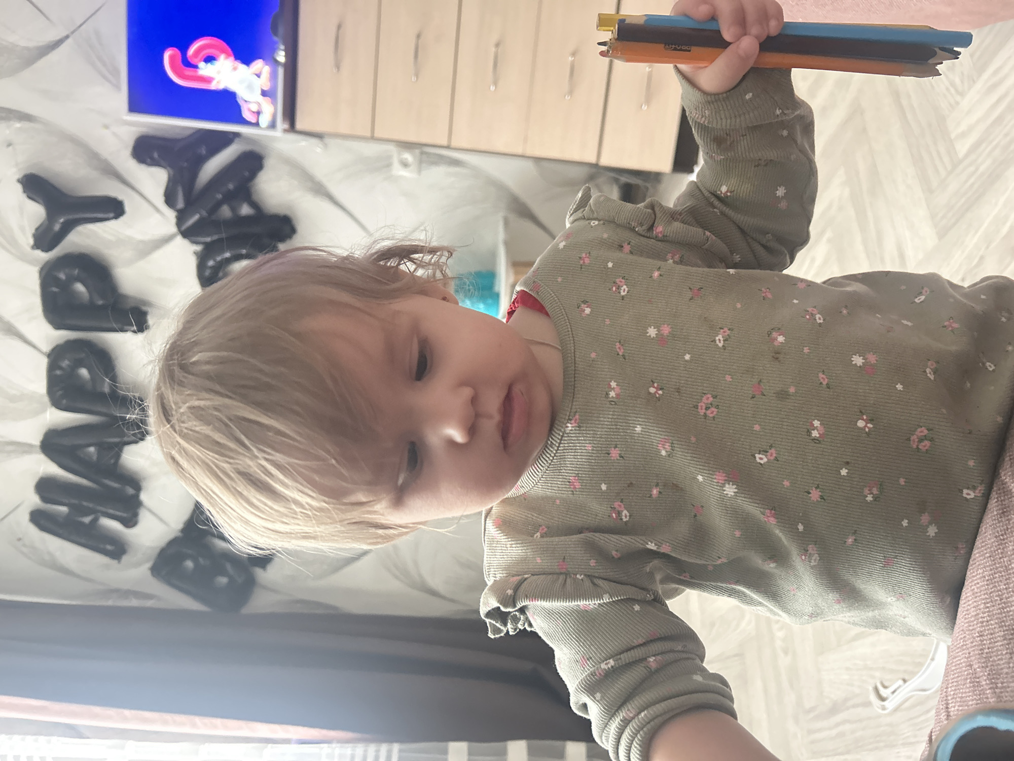 A young child is holding a set of colored pencils in an indoor setting. The child has light blonde hair and is wearing a green, floral-patterned long-sleeved shirt. Behind the child is a wall with black "HAPPY" balloons, a television showing a cartoon, and a dresser. The location is possibly Chaikovsky, Russia.