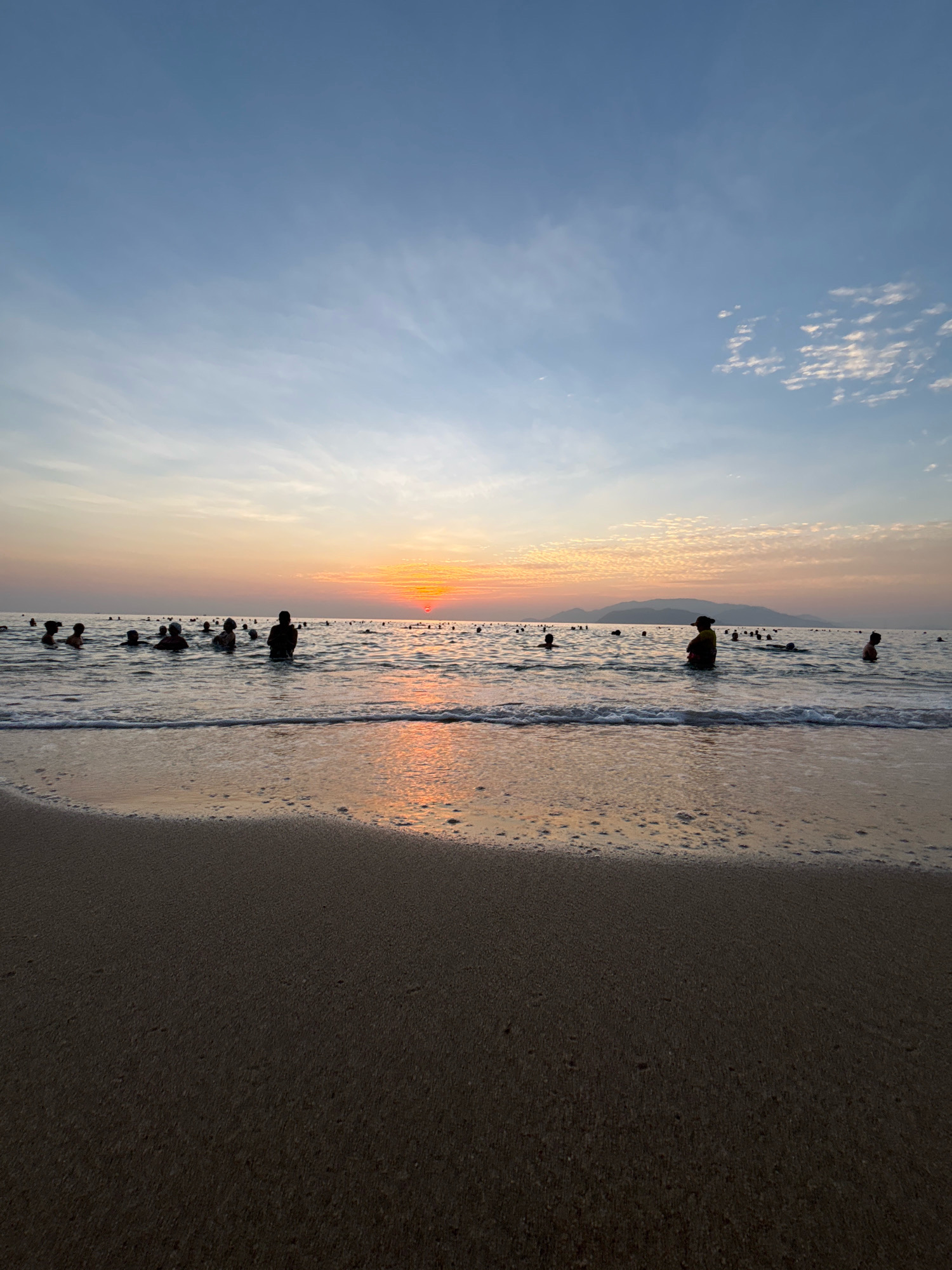 The image shows a beach scene at sunset, likely in Nha Trang, Vietnam. The sky is a gradient of blue and orange, with scattered white clouds. The sun is setting over the horizon, casting a warm glow on the water and the sky. Several people are wading and swimming in the ocean, their silhouettes visible against the colorful sunset. The water is calm, with small waves lapping at the sandy shore. The foreground is dominated by the wet sand of the beach, reflecting the colors of the sunset. In the distance, a line of mountains is visible against the horizon.