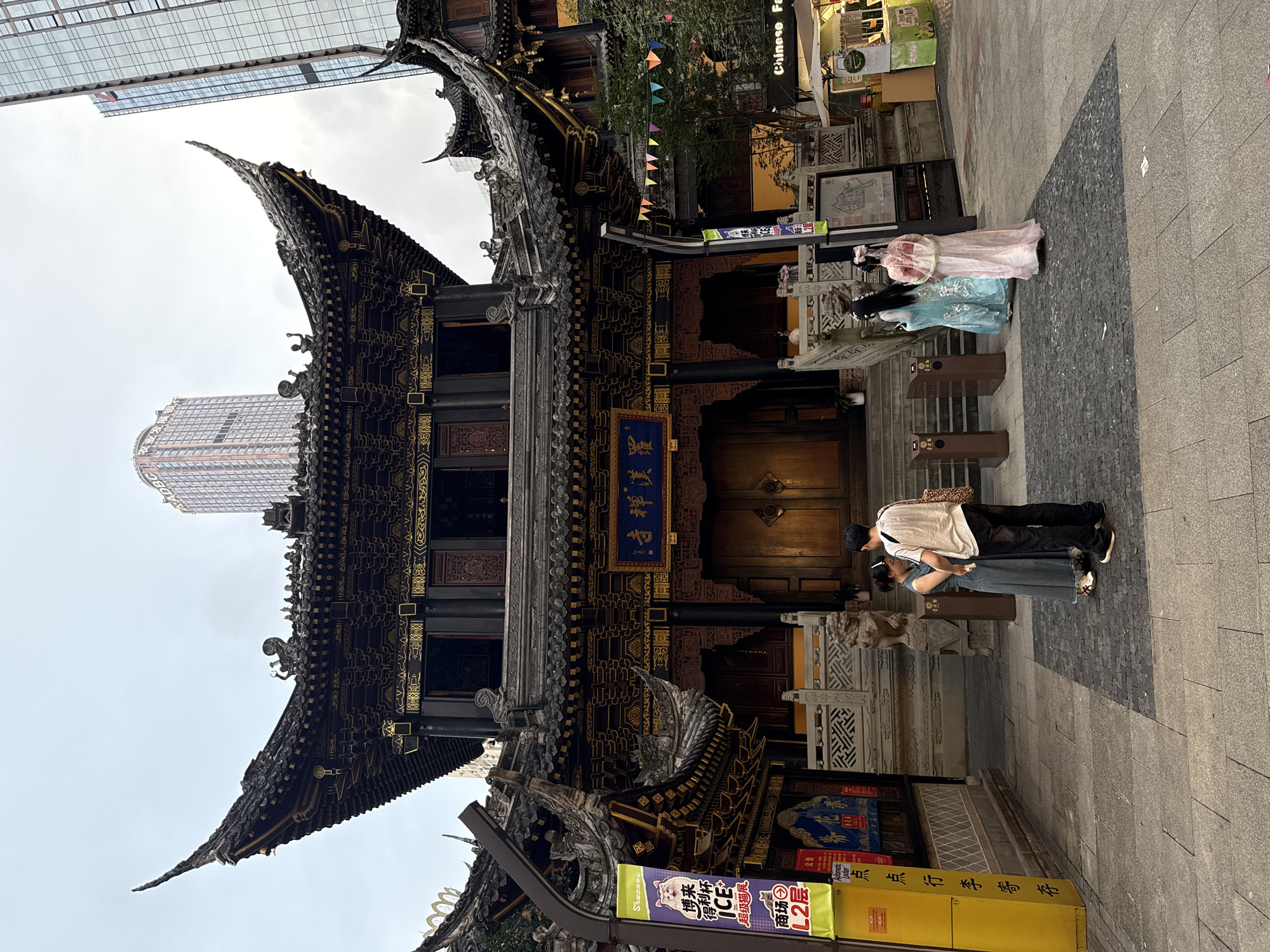 The image depicts the exterior of the Chenghuang Temple in Yuzhong District, China, with a clear view of its traditional Chinese architecture. The temple features ornate, dark-tiled roofs with upturned eaves, adorned with intricate golden patterns. The main building has large, golden double doors and is flanked by smaller structures with similar architectural styles. In front of the temple, a stone staircase leads up to the entrance, guarded by stone lion statues.
Several individuals are present in the foreground. On the left, a couple, a man and a woman, are standing together, facing the temple. The man is wearing a white t-shirt and dark pants, while the woman is in a light blue, sleeveless dress. To the right of the couple, two women are standing, both dressed in traditional Chinese attire. One is wearing a light blue gown, and the other a pink and white outfit, with their backs partially turned to the viewer.
The surrounding environment includes a paved courtyard with a mixture of grey and dark patterned tiles. To the left of the temple, a tall yellow pillar with Chinese characters and promotional banners is visible. In the background, modern skyscrapers rise above the temple's roofline, indicating the juxtaposition of historical and contemporary architecture in the urban setting. Small colorful triangular flags are strung across the area in front of the temple, adding a festive touch. Various signs and storefronts, including one labeled "Chinese Fashi," are also visible on the right side of the image, suggesting a commercial area adjacent to the temple. The sky is overcast, suggesting a cloudy day.