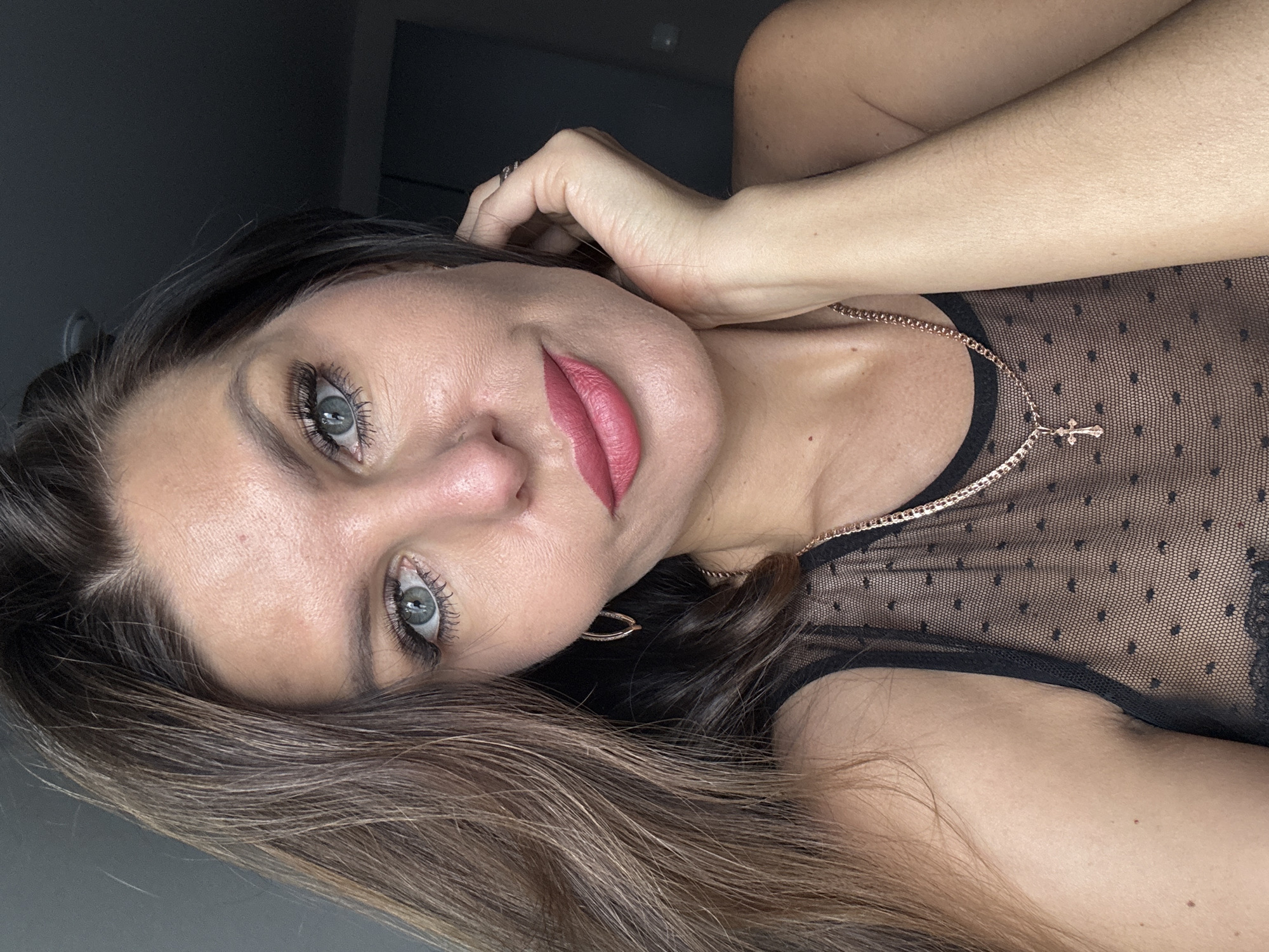 A woman with light skin, long brown hair, and blue eyes is facing the camera. She is wearing a black sheer top with small polka dots and a gold chain with a cross pendant. She is also wearing lipstick and has mascara on her eyelashes. She is touching her hair with her right hand, where a ring is visible. She appears to be indoors, with a gray wall behind her. The location and city are not discernable.