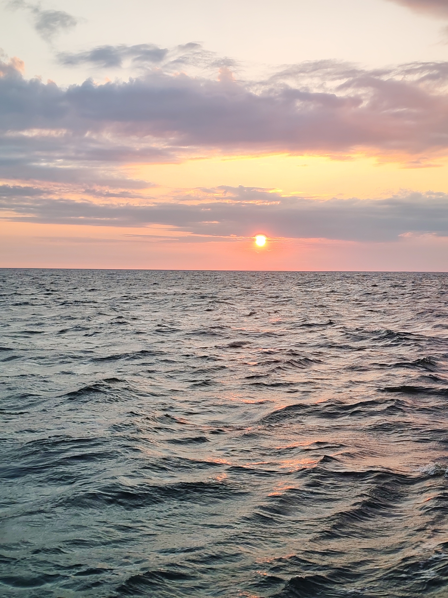 The image depicts a serene seascape at sunset. The sky is a canvas of soft hues, transitioning from a pale yellow near the horizon to delicate pinks and purples, with scattered, wispy clouds. The sun, a bright, orange orb, is partially obscured by clouds, casting a warm glow across the sky and reflecting shimmering streaks on the water's surface. The sea itself is calm, with gentle ripples creating a textured, dark blue expanse that stretches to meet the horizon. The scene is devoid of any human presence or man-made structures, conveying a sense of natural tranquility. There is no information available regarding the specific location within Unknown, Russia.