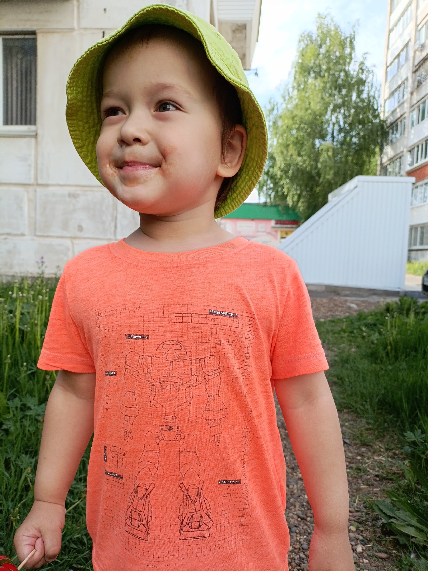 A young child stands outdoors on a pathway lined with grass and weeds. He wears a bright orange t-shirt with a schematic design of a robot or mecha on it. He also wears a green sun hat. There's evidence of food or chocolate smeared around his mouth. A small object on a stick is visible in his hand. A low white building or structure is visible behind him, along with parts of a taller multi-story building. Based on geolocation, this image may be located in the city of Chaikovsky, Russia.