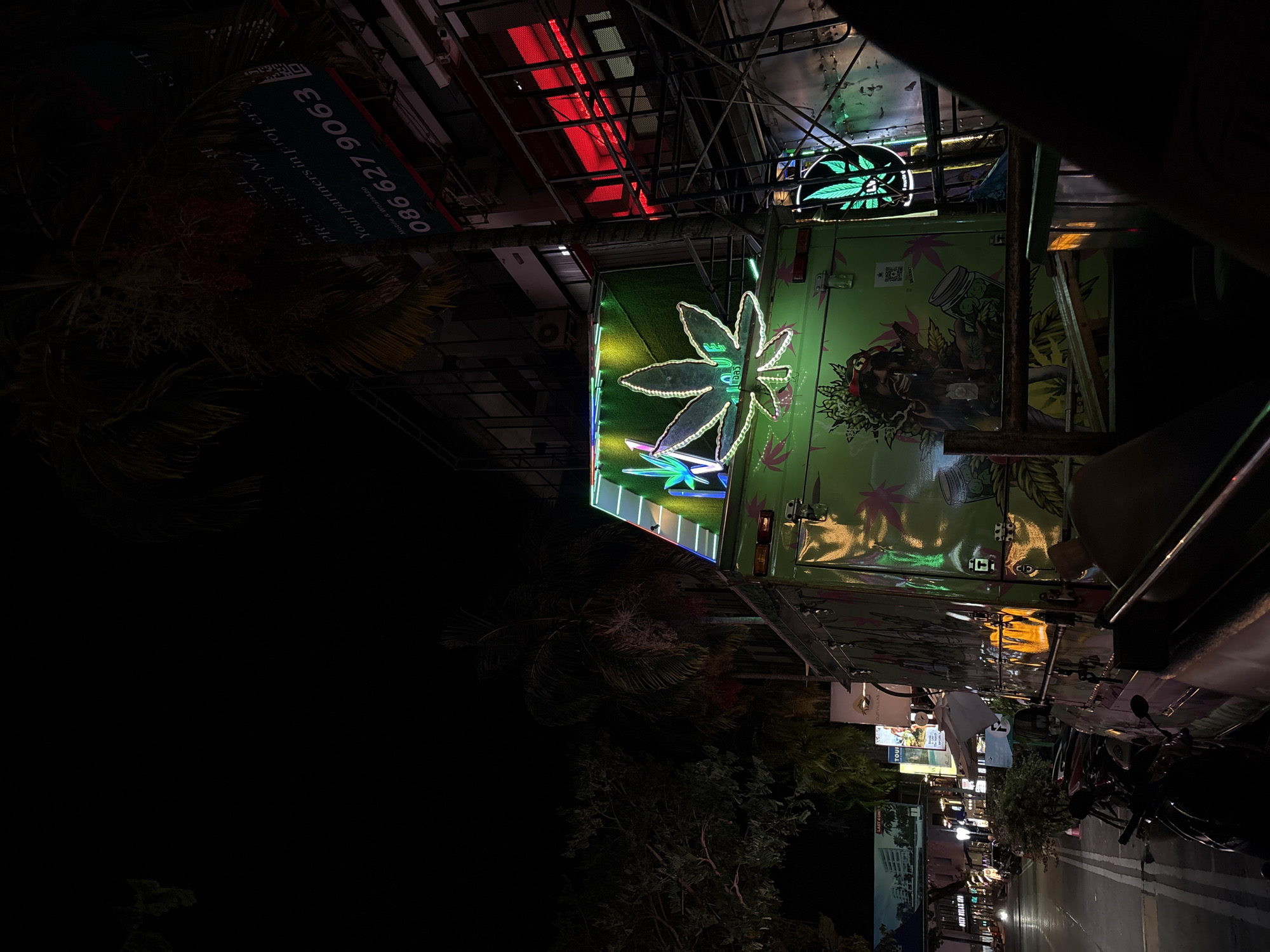 The image shows a nighttime street scene in Choeng Thale, Thailand. A brightly lit green food truck with cannabis leaf imagery is parked along the side of the road. The truck features neon signs in the shape of cannabis leaves, one prominent on its roof and another on its side. The side of the truck is decorated with a mural depicting a stylized human figure holding a jar filled with cannabis. Several neon lights and signs illuminate the scene, including a red neon sign on a building in the background. Palm trees and other vegetation are visible, as is a street lined with buildings and parked motorcycles. The street is wet, suggesting recent rain, and reflects the light from the signs. A portion of a person's arm and shoulder is visible in the foreground, obscuring part of the food truck. A sign on a building in the background displays a phone number: "086 627 9063". The overall atmosphere is vibrant and commercial, illuminated by artificial light against the dark night sky.