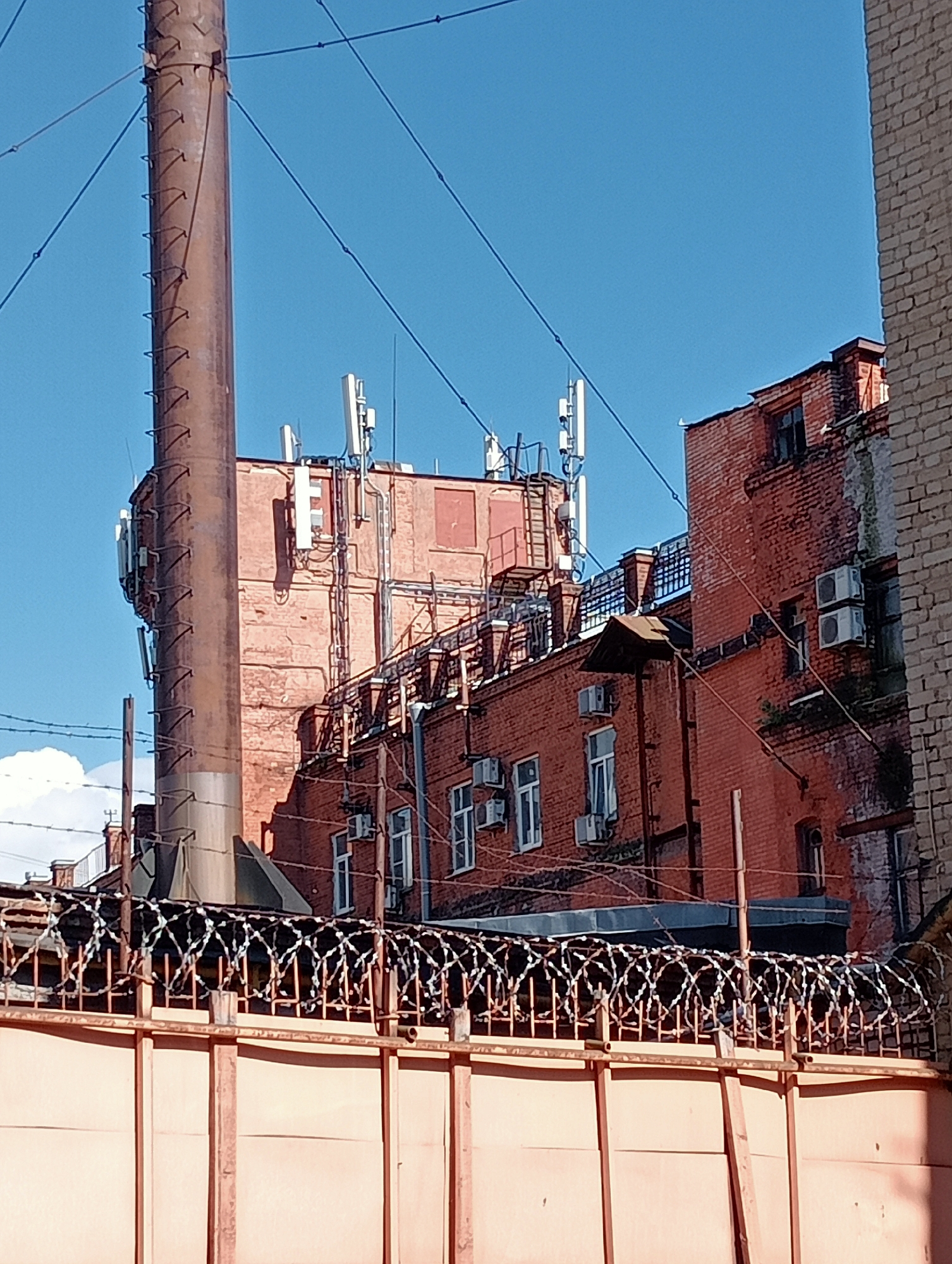 The image depicts an industrial or urban scene, likely in Saint Petersburg, Russia, characterized by red brick buildings and a tall, weathered smokestack. The sky is a clear, bright blue, suggesting a sunny day, possibly midday given the strong shadows.
Dominating the left side of the frame is the large, rusty smokestack, which has a ladder-like structure attached to its side. Several communication antennas, likely for cellular networks, are mounted on the rooftops of the red brick buildings behind the smokestack. These buildings appear to be multiple stories high and show signs of age, with some areas of peeling paint or discolored brickwork. Windows are visible on the buildings, and some have air conditioning units installed.
In the foreground, a metal fence topped with barbed wire stretches across the image, creating a sense of confinement or security. The fence and the wall below it are a muted orange or brown color. The overall atmosphere is one of an urban industrial area, possibly a former factory or a complex of utilitarian buildings. The clear sky and sunlight highlight the textures of the brick and metal. No people are visible in the image, and there is no discernible text. The scene is static, with no specific activity or event taking place.