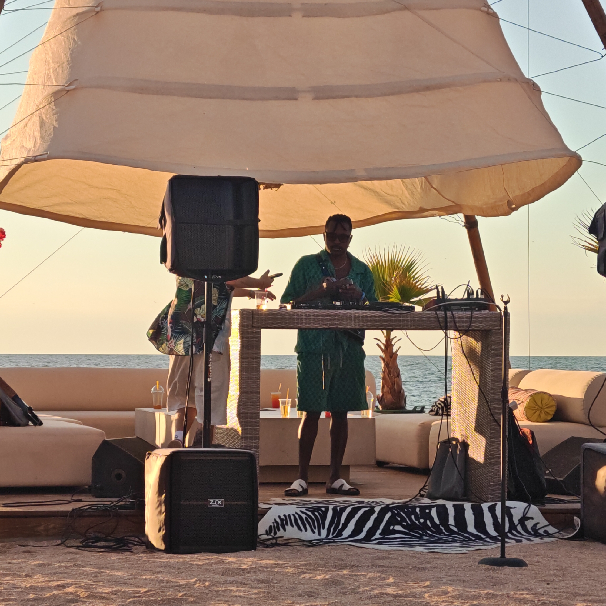 The image depicts an outdoor scene near a body of water, likely the sea, with a light-colored canopy overhead. Two individuals are present. One person, wearing sunglasses, a green patterned shirt, and shorts, stands behind a wicker table with DJ equipment. They appear to be handling some items. Another person, partially visible on the left, wears a patterned shirt and light-colored pants, and holds a cup with a straw. The environment includes a sandy foreground with a zebra-patterned rug and two large black speakers. Behind the DJ table, there are lounge chairs and a palm tree. The water is visible in the background, suggesting a coastal or beachside location. The lighting suggests it is late afternoon or early evening, with warm, golden light illuminating the scene. There is no specific indication of the city of Щёлкино, Ukraine in the image.