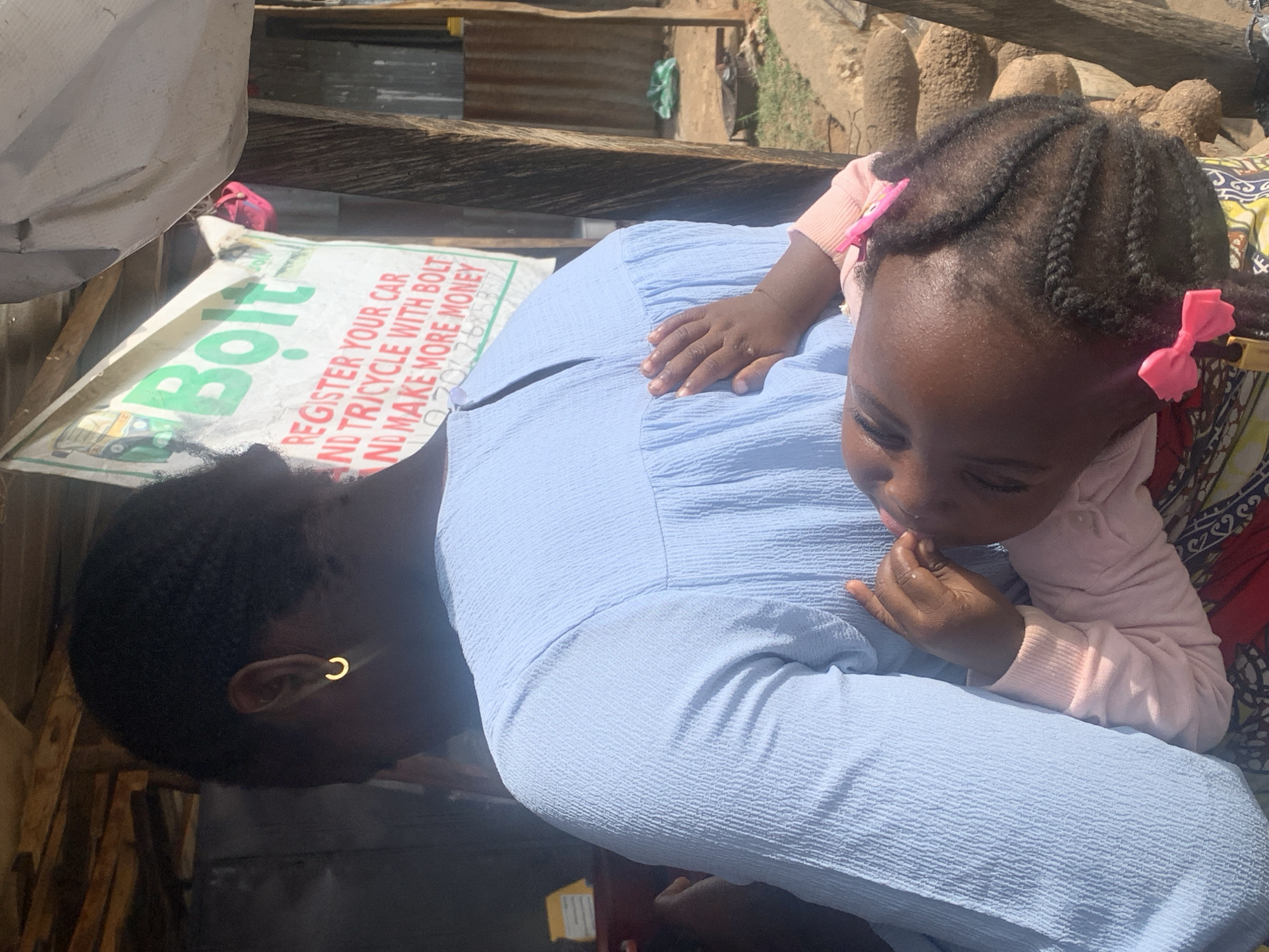 A woman and a young child are depicted in an outdoor setting, likely within a residential area in Jos, Nigeria. The woman, positioned on the left side of the frame, has short, dark hair and wears a simple gold hoop earring. She is dressed in a light blue, possibly linen or textured fabric dress. The child, adorned with two pink bows in their braided hair, rests against the woman. The child wears a light pink sweater. The background includes what appears to be a Bolt advertisement, suggesting the presence of ride-hailing services in the area. The immediate environment shows signs of modest or informal housing, with corrugated metal sheets, wood structures, and potentially natural fencing elements visible. The setting suggests a low-income neighborhood in Jos, characterized by its informal construction and potential resource constraints.