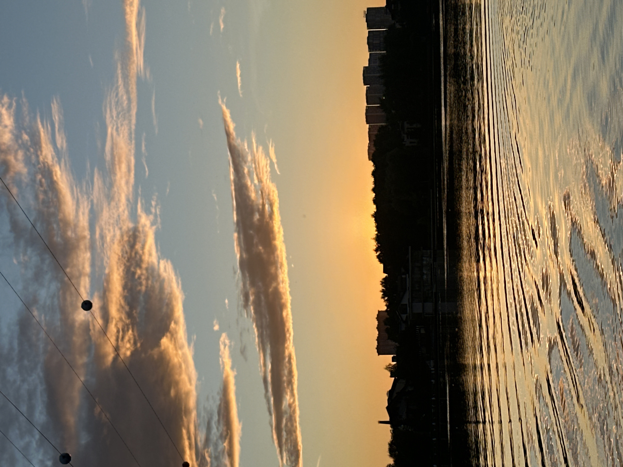 The image depicts a waterfront scene at sunset in Myakinino, Russia. The sky is a gradient of pale blue and warm orange, with long, wispy clouds tinged with golden light. In the foreground, dark wires with evenly spaced spherical objects stretch across the frame, suggesting utility lines or decorative lighting. To the right, the water reflects the sky, creating shimmering patterns of light and shadow. The surface of the water is disturbed by ripples, giving it a textured appearance. Along the horizon, a silhouetted treeline forms a dark band, behind which rise the stark shapes of several apartment buildings. The warm hues of the sunset and the calm water suggest a peaceful atmosphere. The image is captured in landscape orientation, but the elements are presented vertically as if the camera was rotated 90 degrees.