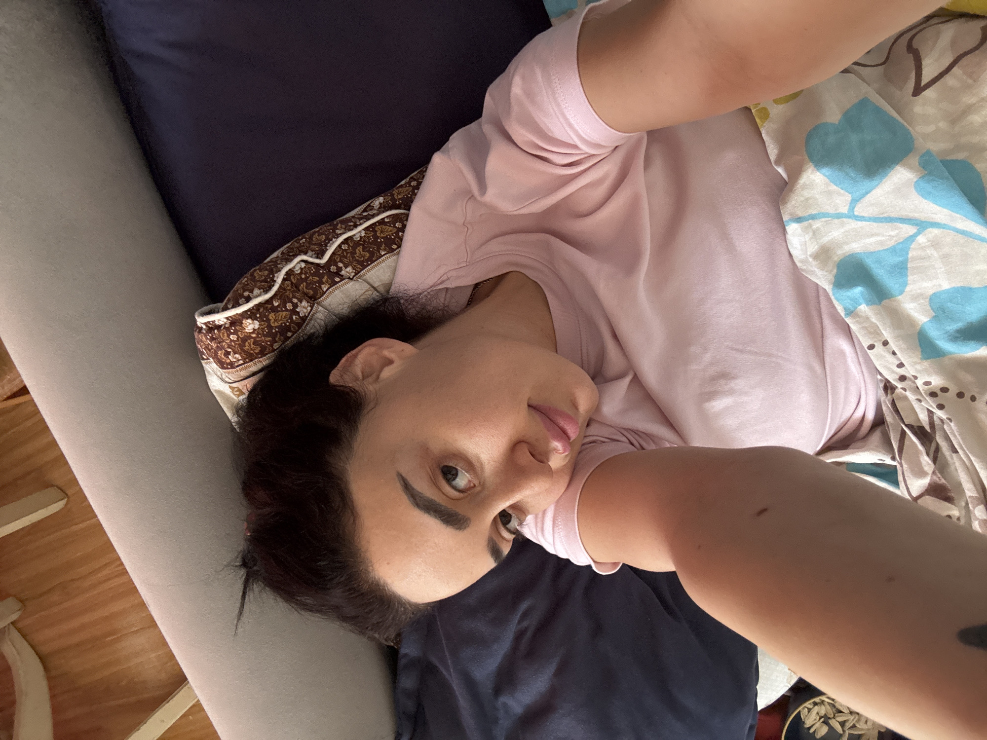A person lies in bed, taking a selfie. The person has dark hair and is wearing a pink t-shirt. The bed has pillows with floral and patterned designs. The city of Tbilisi, Georgia is not visually confirmed.