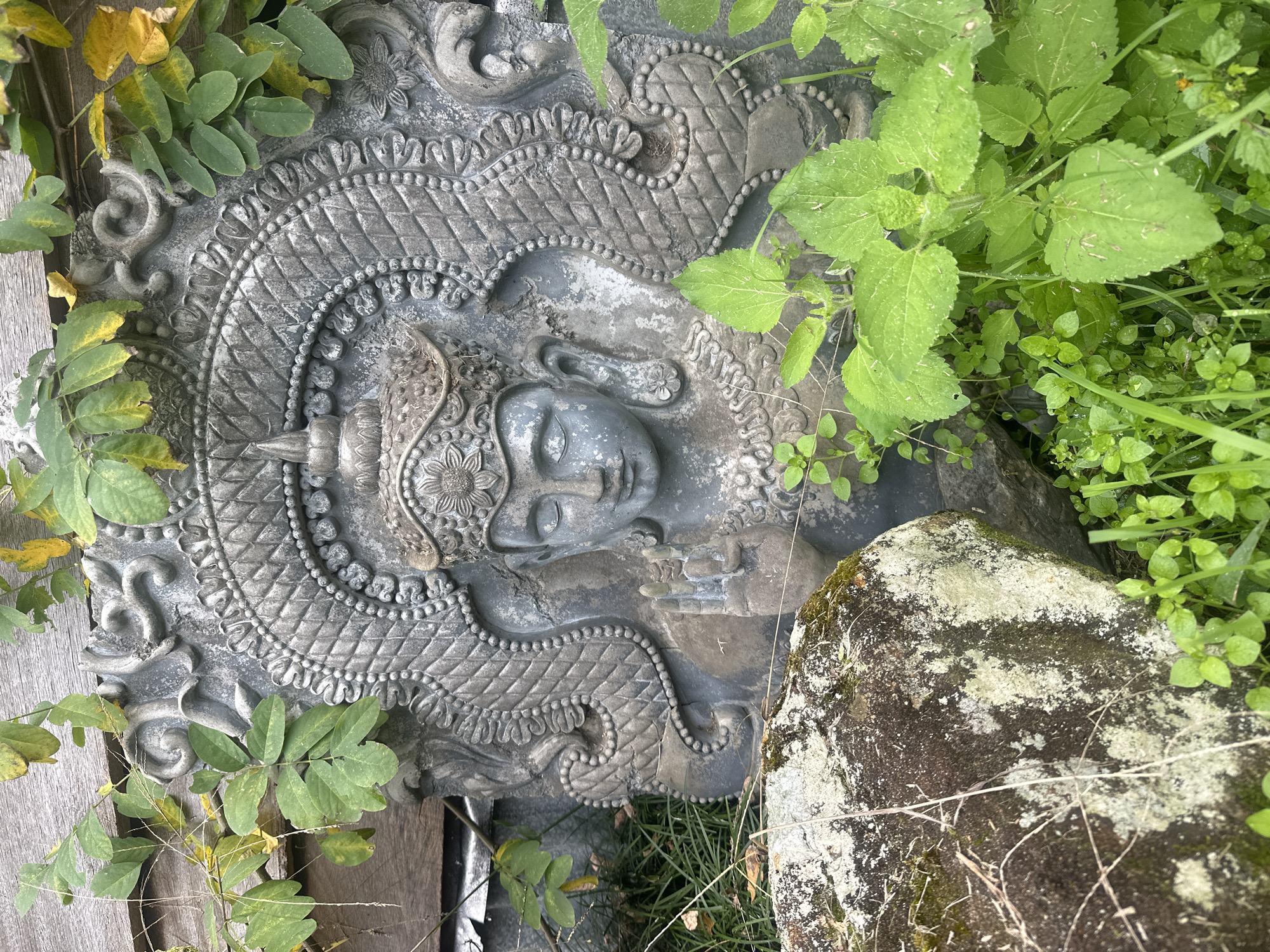 An ornate, weathered stone carving of a deity is partially obscured by lush green foliage and a large, moss-covered rock. The carving, rendered in a gray stone, depicts a figure with closed eyes, a headdress, and intricate decorative elements surrounding its face, including what appear to be stylized dragon-like motifs. The figure's right hand is raised with palm facing outward, possibly in a gesture of blessing or protection. The carving is set against a backdrop of weathered wooden planks. The scene is situated outdoors, with the vegetation and natural elements suggesting it is in a garden or natural setting. The specific location in the city of Thora, Australia, is not visually identifiable from the image.