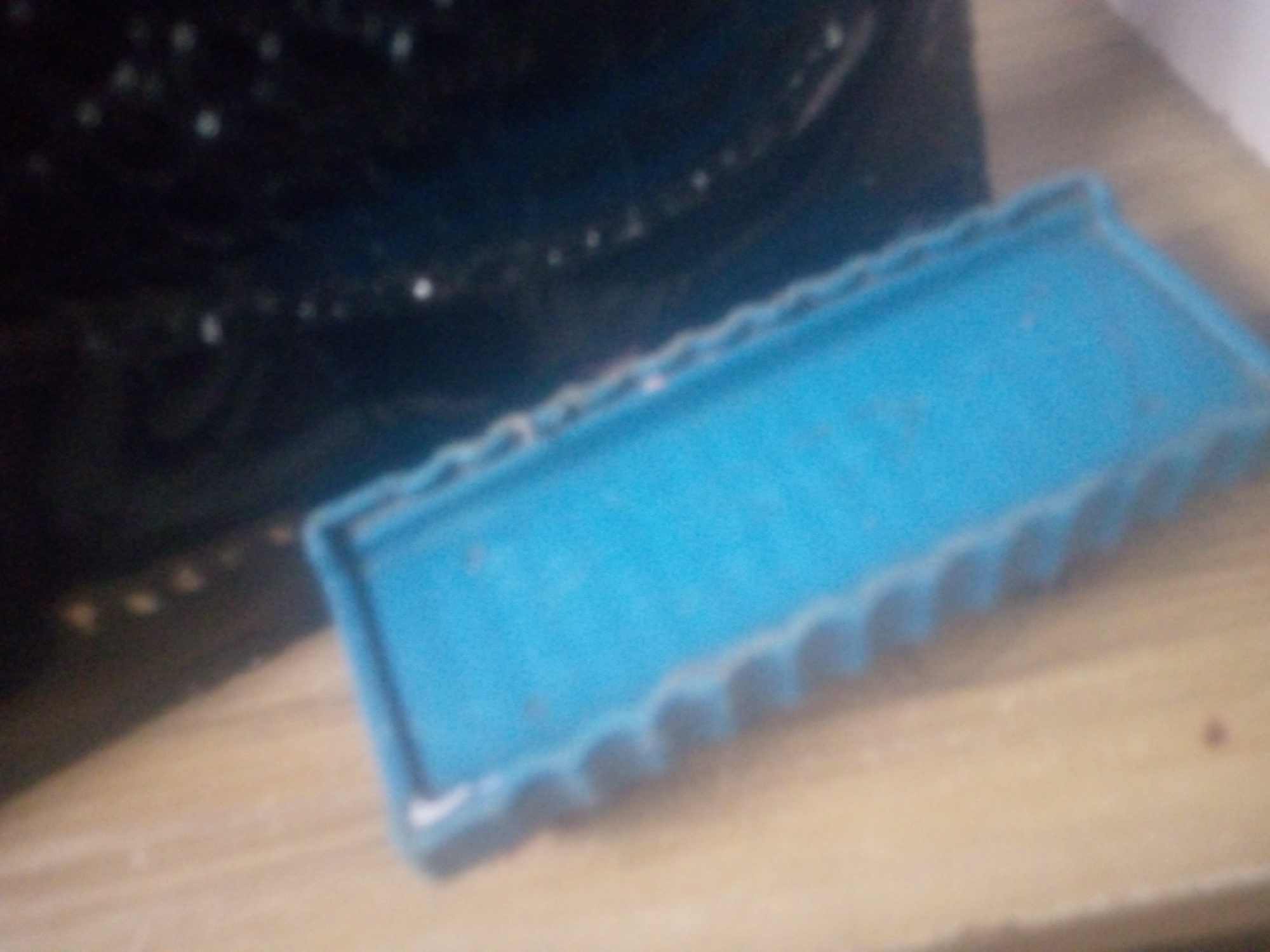 The image shows a close-up, slightly blurred view of a blue object, possibly a plastic container or tray, with a textured edge. The object is resting on a light-colored, wooden surface. Behind the blue object, a dark, possibly black, reflective surface is visible, with several small white or light-colored specks scattered across it. The lighting suggests an indoor setting. The overall composition is focused on the blue object and its immediate surroundings, with no identifiable city, state, or specific location details present.