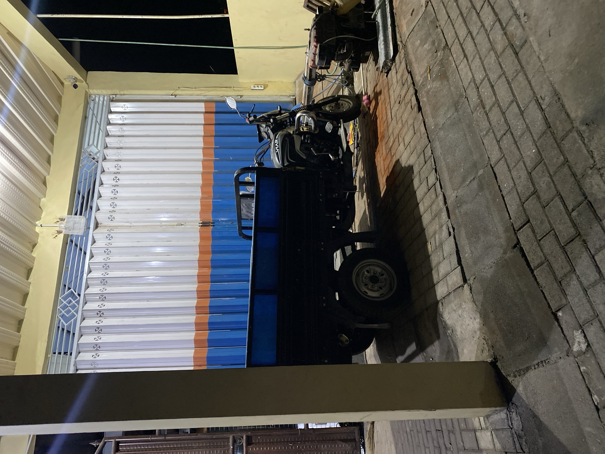 A three-wheeled motorcycle with a cargo bed is parked in front of a closed garage with a white, orange, and blue striped corrugated metal door. To the right of the motorcycle, an engine sits on the ground, connected to a hose. The scene is illuminated by artificial light, casting shadows on the brick-paved ground. The image appears to have been taken at night in a somewhat industrial or commercial area. The location is Kuta Selatan, Indonesia, based on the context provided.