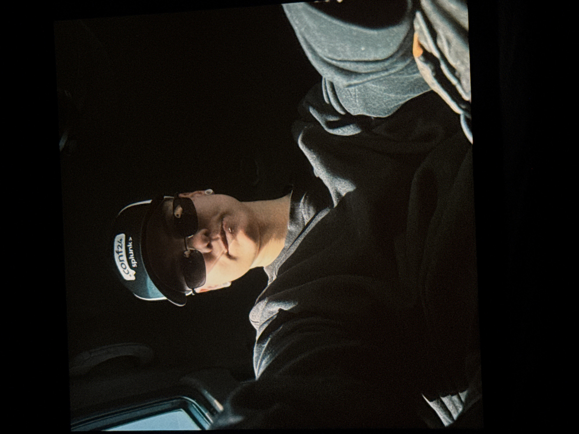 A person wearing a dark baseball cap and sunglasses is seated in the interior of a vehicle. The cap has a logo that reads "conf24 splunk>". The person is wearing a dark gray crew neck sweatshirt. The background is dark, with a small portion of a window visible on the left side of the frame. The lighting suggests this is daytime. The image is a close-up shot, focusing on the person's upper body. There are no discernible elements that place the scene specifically in Irkutsk, Russia.