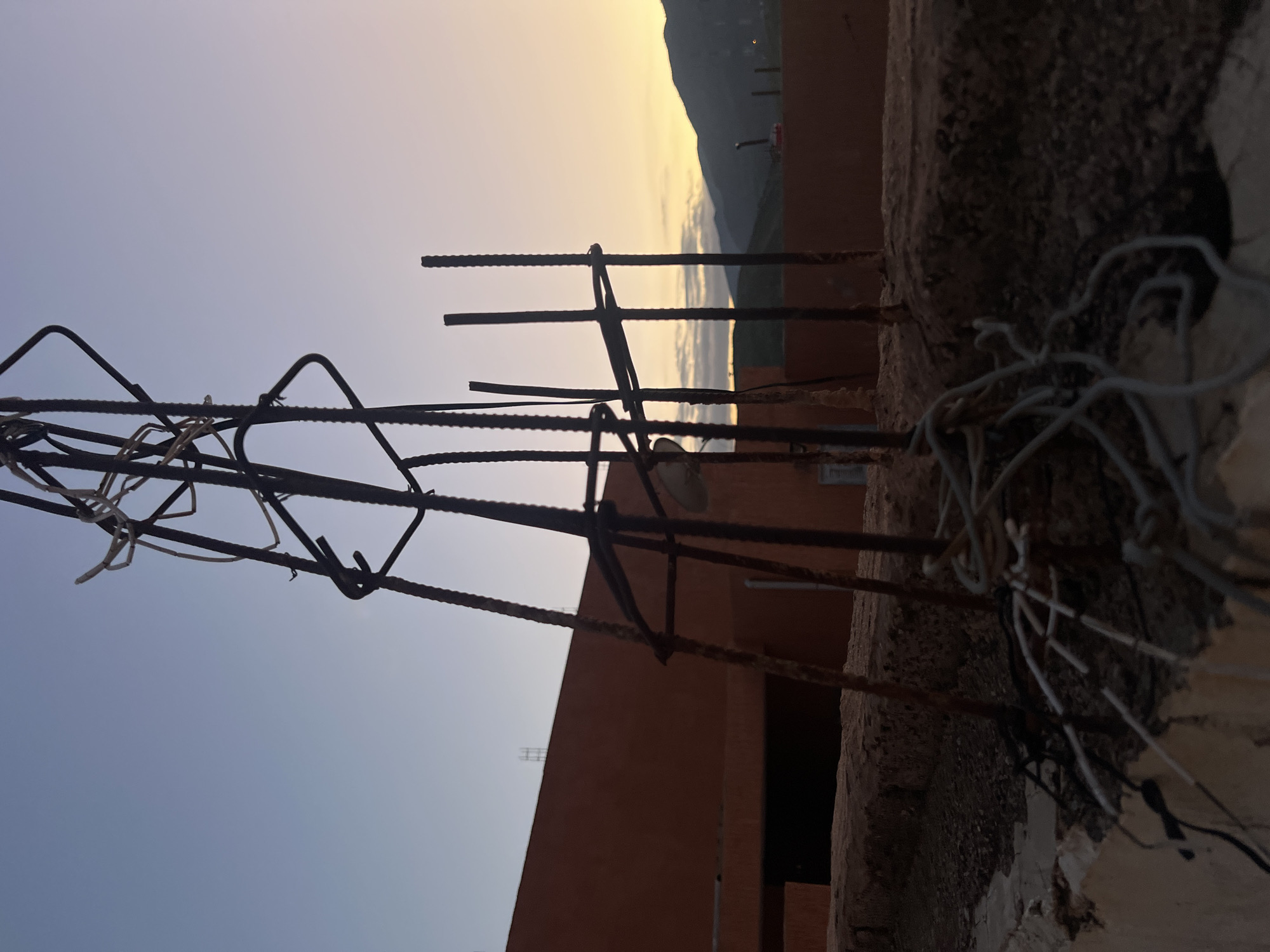 A ground-level shot in Ouaouizert, Morocco, focuses on construction rebar jutting out from a partially constructed brick building. The rebar is rusty and secured with wire, some of which hangs loosely. In the background, a mountain range is visible under a sky transitioning from light blue to a yellow-tinged sunset. The building's exterior is unfinished, revealing raw materials.