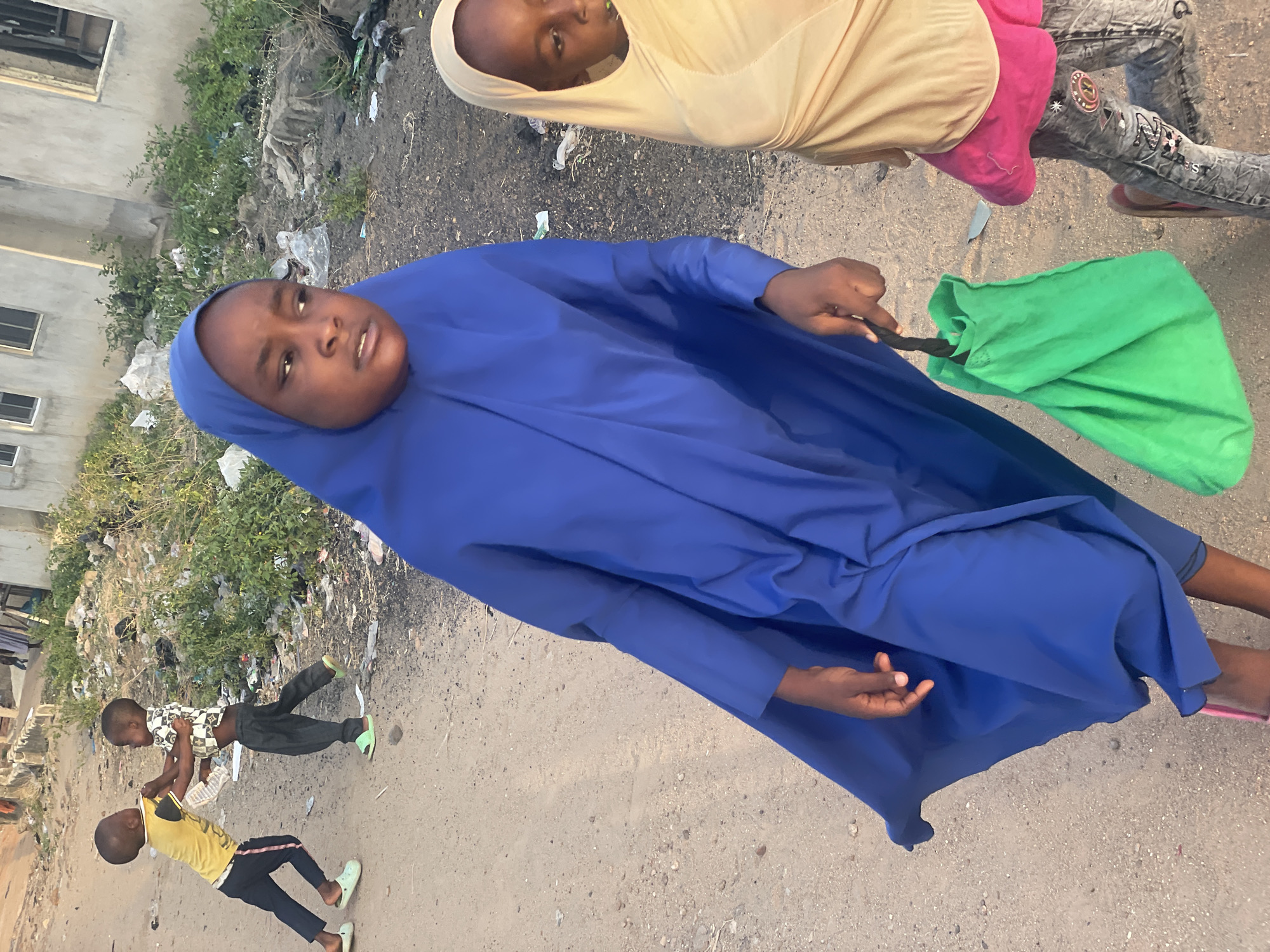 A young girl wearing a royal blue hijab stands on a paved road in what appears to be Lokoja, Nigeria. She holds a green bag in her right hand and looks directly at the camera. To her right, a child in a beige hijab and pink top is also present. In the background, two young boys engage in playful activity. Trash is scattered near a building in the background.