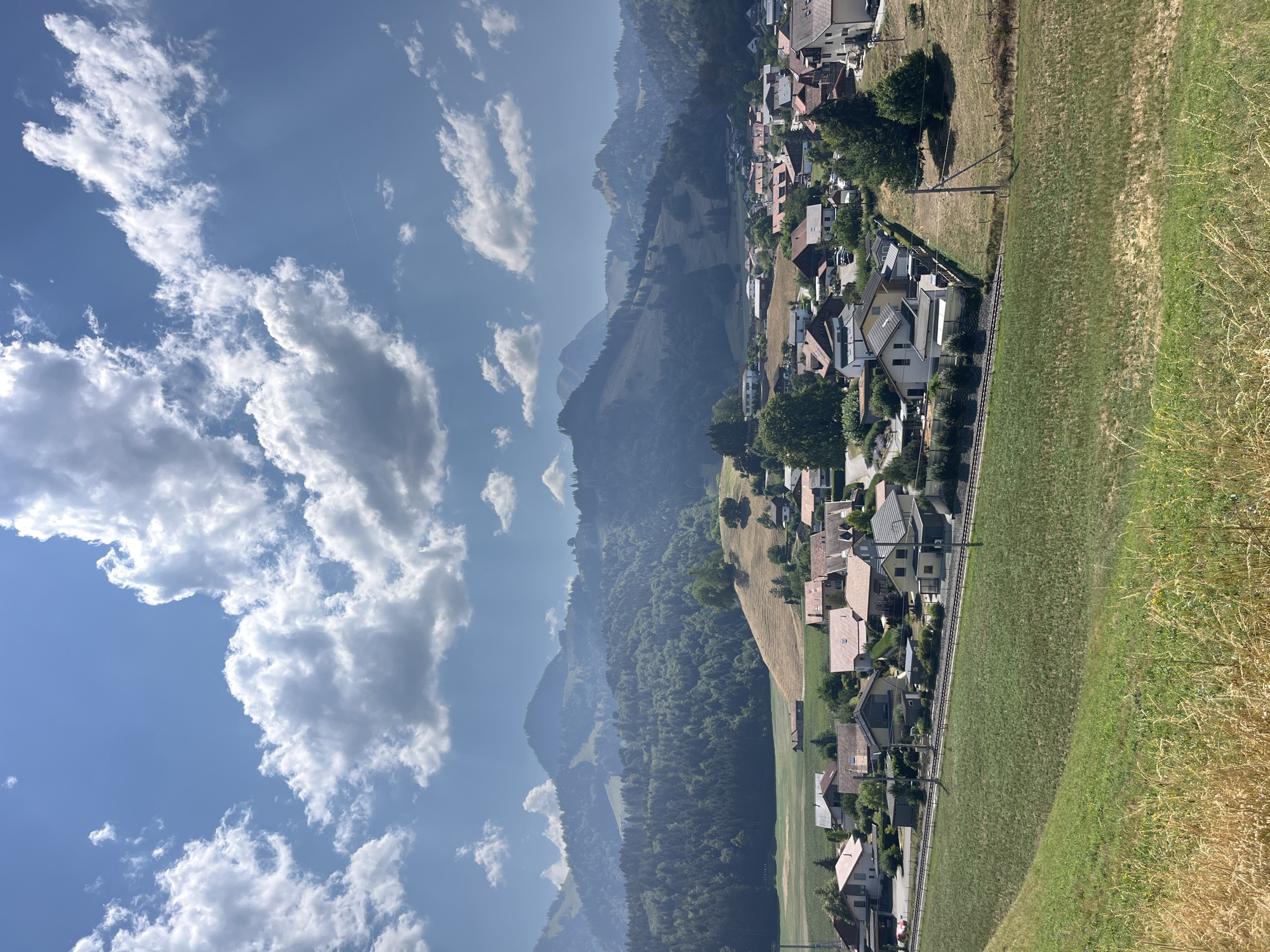 The image depicts a scenic view of the village of Pringy, Switzerland, nestled amidst rolling hills and mountains. The foreground features a lush green grassy field, transitioning to a dry, golden field with tall grasses. A railway track runs horizontally across the middle ground, with several houses and buildings situated on either side. These structures are varied in size and style, with many featuring red-tiled roofs. Beyond the village, the landscape ascends into tree-covered hills and culminates in distant, hazy mountains. The sky is a vibrant blue, dotted with fluffy white cumulus clouds, some of which are illuminated by sunlight, casting shadows on the mountainside. The overall impression is one of a serene, rural setting under a bright, sunny sky.
