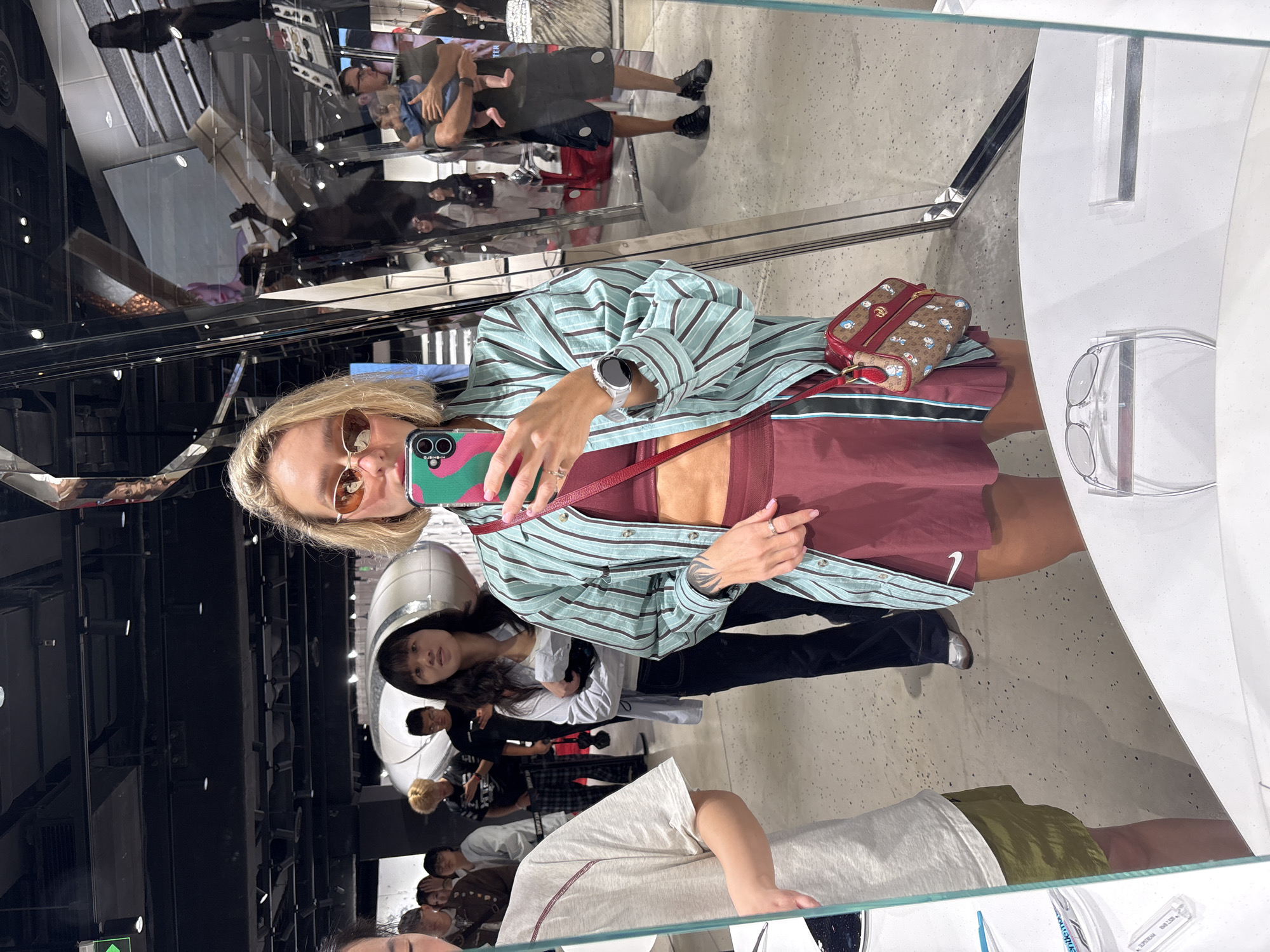 A young woman with blonde hair and sunglasses is taking a mirror selfie in what appears to be a retail store in Shanghai, China. She is wearing a teal and dark blue striped button-up shirt over a maroon sports bra and a matching maroon pleated skirt. She has a small brown and red patterned handbag with cartoon characters hanging from her shoulder. Her left wrist is adorned with a silver smartwatch.
In the background, reflected in the mirror, are other shoppers and store displays. To her left, a woman with dark hair is visible, also looking into the mirror. Further back, a man is holding a baby. The store environment features dark ceilings with black track lighting and a reflective, metallic structure in the mid-ground. The floor is a speckled grey concrete. In the foreground, a white counter with what appears to be eyewear displays is visible.