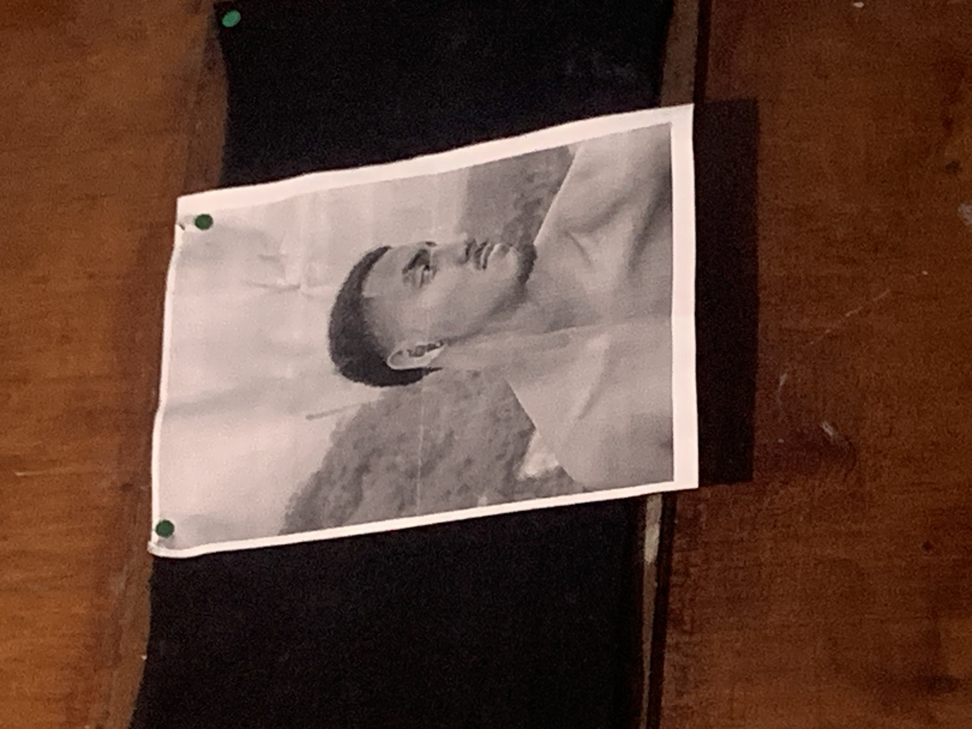 A black and white photograph is prominently displayed, pinned horizontally to a vertical surface. The photograph depicts a young man, likely in his late teens or early twenties, with short, dark hair. He is shown in a three-quarter profile, looking upwards with a neutral or contemplative expression. His upper torso is visible, suggesting he is shirtless or wearing light clothing.
The photograph is held in place by three small, green thumbtacks: one in each of the top corners and one in the bottom-left corner. Directly behind the photograph is a strip of dark, possibly fabric or felt, material. This dark material is framed by a reddish-brown wooden surface, which forms the surrounding wall or paneling. The overall setting appears to be an indoor space, characterized by simple, possibly rustic, materials.
There are no discernible indications of the time of day, weather, or specific location cues within the image itself. No text is visible. The scene portrays a personal display of a photograph within an unadorned interior environment, set in Santa Ana, Venezuela as per the contextual information.