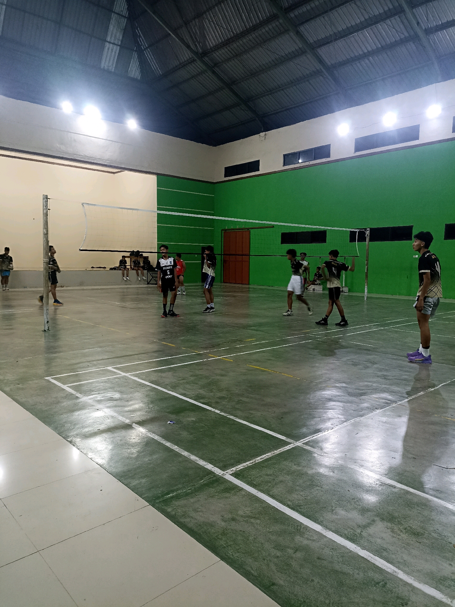 The image depicts an indoor volleyball court in Sumberharjo, Indonesia. Several young men, likely athletes, are present on the court, some actively engaged in a game while others observe from the sidelines. The court is marked with white lines on a green floor. A volleyball net is set up across the court. The indoor space features a metal roof with industrial lighting fixtures illuminating the area. The walls are painted in green and white. The overall scene captures a moment of athletic activity in a communal indoor sports facility.