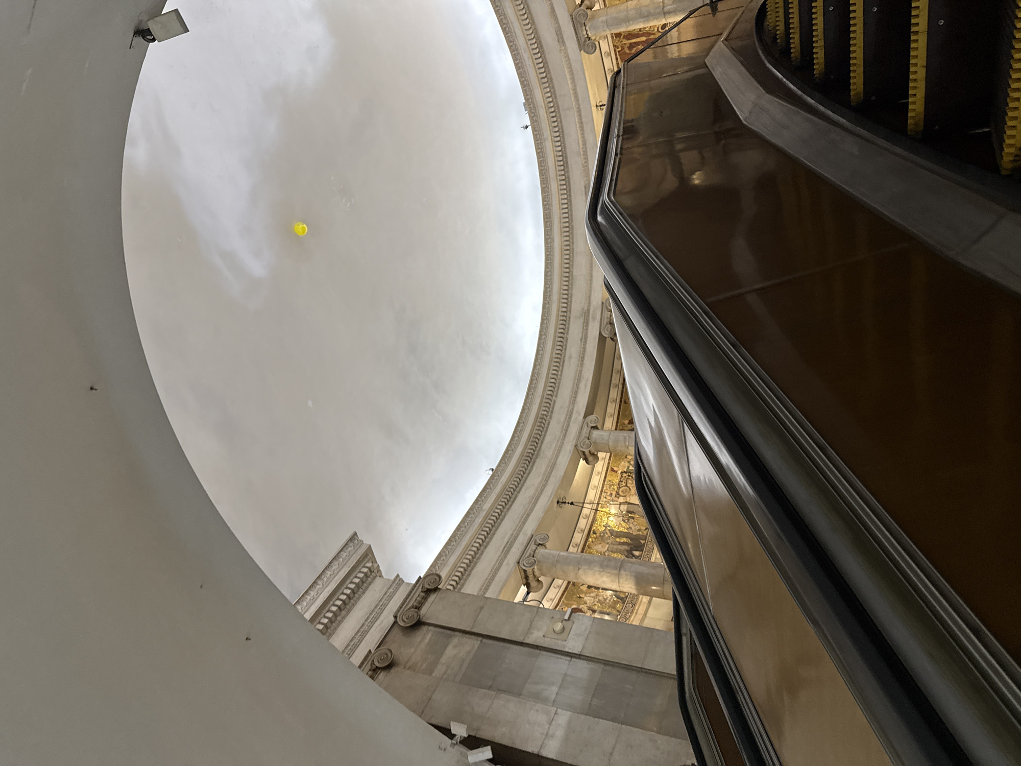 The image captures an interior view of a grand metro station in Moscow, Russia. A wide escalator with dark brown sides and bright yellow steps dominates the foreground, angled upwards towards the left. Above the escalator, a high, domed ceiling is visible, adorned with classical architectural elements including fluted columns with Corinthian capitals and ornate decorative moldings. Faint murals or mosaics can be seen on the walls behind the columns, depicting figures and scenes. In the upper portion of the domed ceiling, a single, small, bright yellow balloon floats against the pale, cloudy sky depicted on the ceiling. The lighting appears to be artificial, casting a soft glow on the opulent surroundings. The overall impression is one of grandeur and architectural splendor characteristic of many Moscow Metro stations.