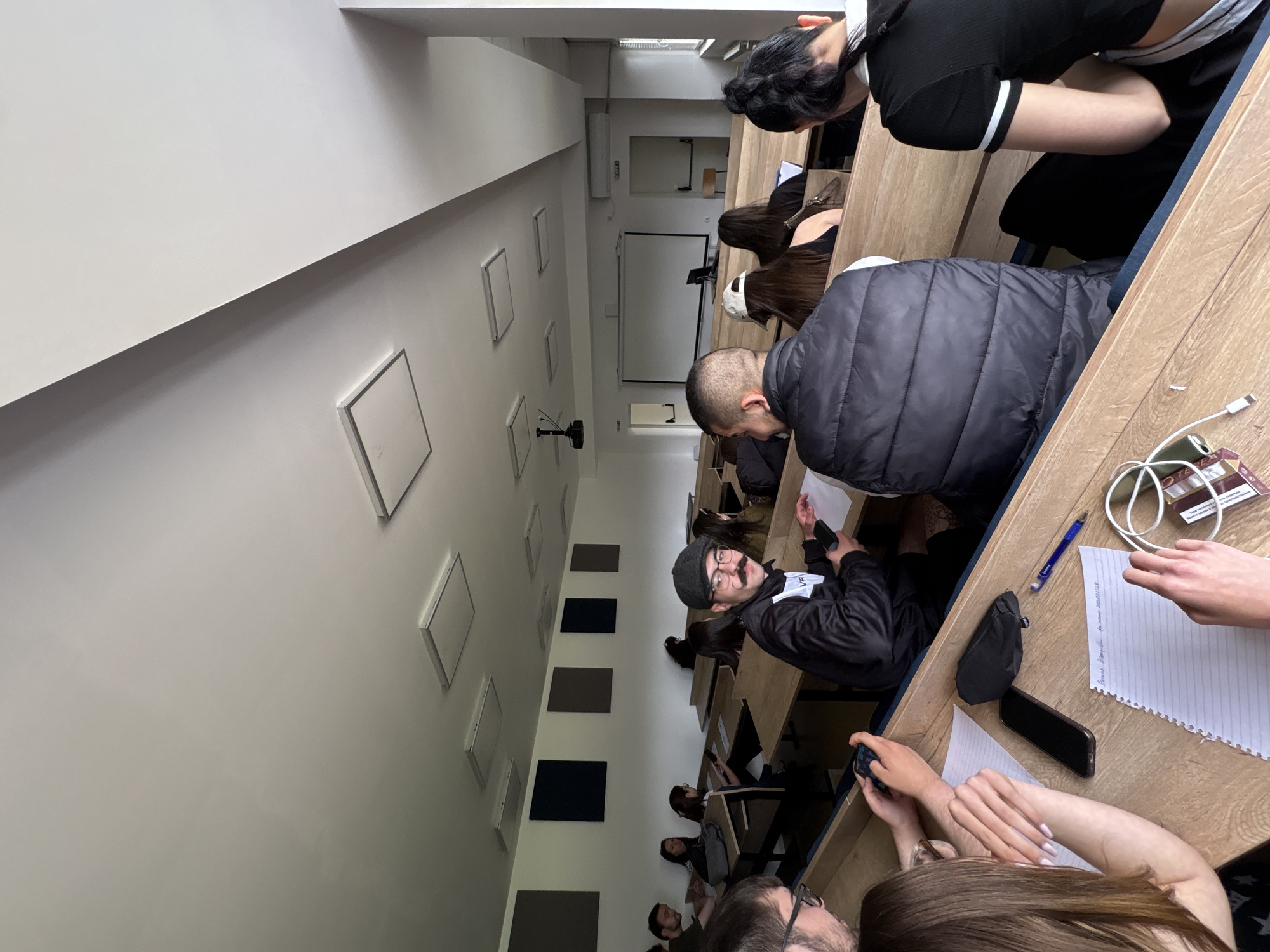 A medium shot captures an elevated perspective inside a lecture hall, likely at the American University in Bulgaria, Blagoevgrad. Students are seated at tiered, wooden desks, some writing on lined paper with pens, others using smartphones. Cables, cigarettes, and a wallet are also on a desk. The wall is painted white with evenly spaced, grey and black square panels. A projection screen is visible at the front.