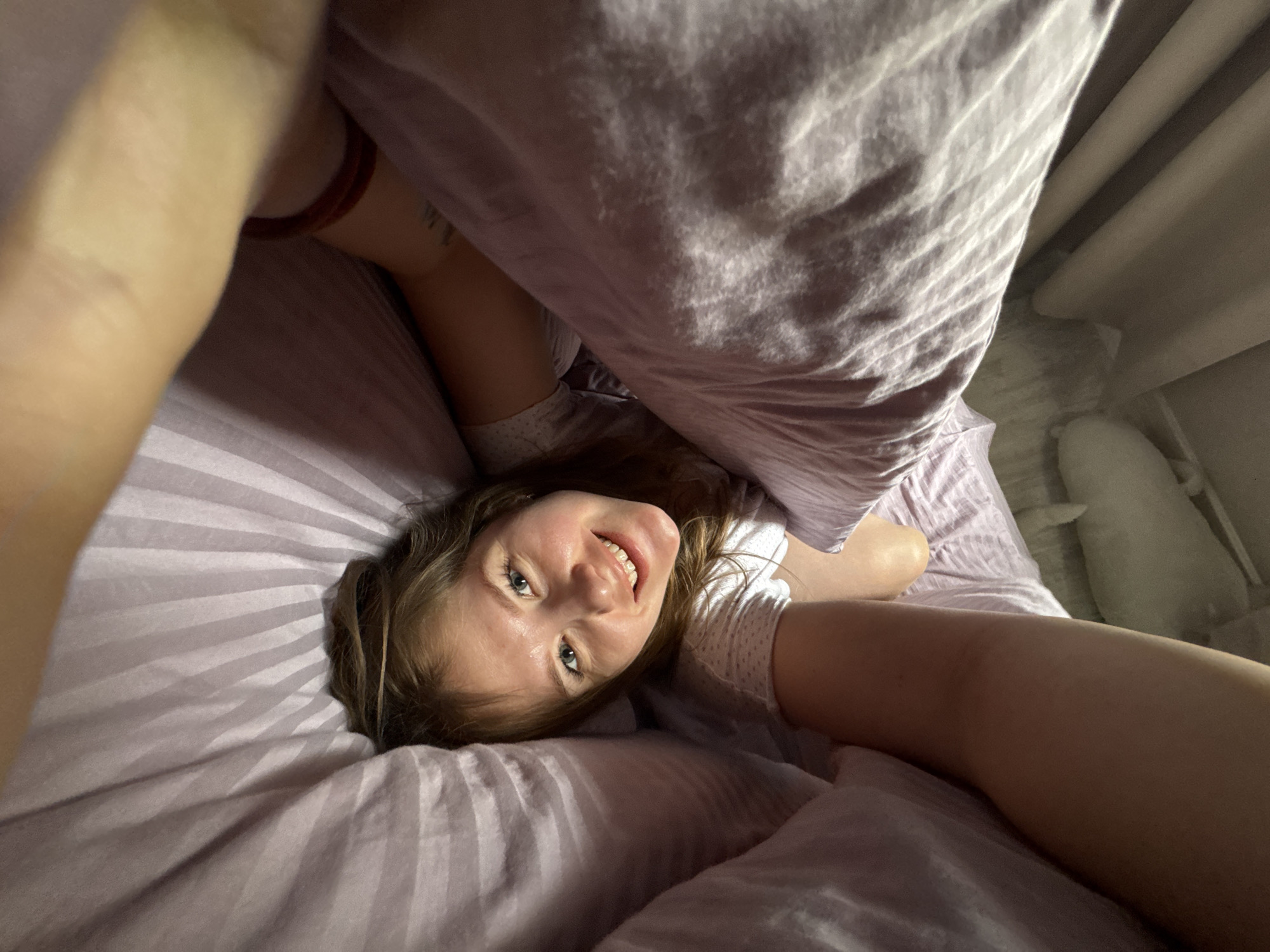 A young woman with long, light brown hair and blue eyes is lying in bed under a light purple striped duvet cover. She is smiling at the camera, which she is holding up to take a selfie. She is wearing a white t-shirt. She is partially covered by the duvet, which is pulled up around her. In the background, there are light-colored curtains and a white pillow. The photo appears to be taken indoors, possibly in a bedroom. There is no indication of the city of Gornyi or Dvurechenskoe S/P in Russia.