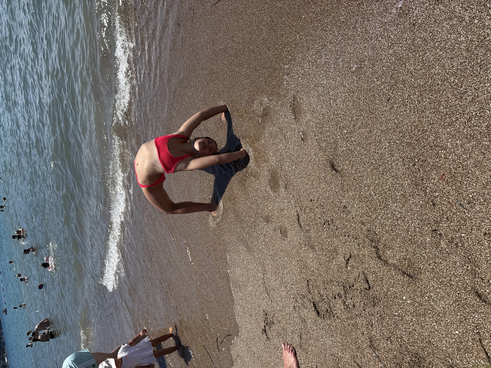 The image shows a beach scene at Budva, Montenegro, on a sunny day. The main subject is a young woman in a red bikini performing a backbend on the sandy shore, with the water lapping at the edge. Her arms are extended, and she appears to be smiling. Her shadow is cast on the sand.
In the background, the blue water of the sea is visible, with several other people swimming and wading. Some are further out in the water, while others are closer to the shore. The sand is coarse and speckled with small pebbles. The sun appears to be high in the sky, suggesting it is midday. The overall mood is relaxed and recreational, typical of a beach holiday. There is no visible text in the image.
