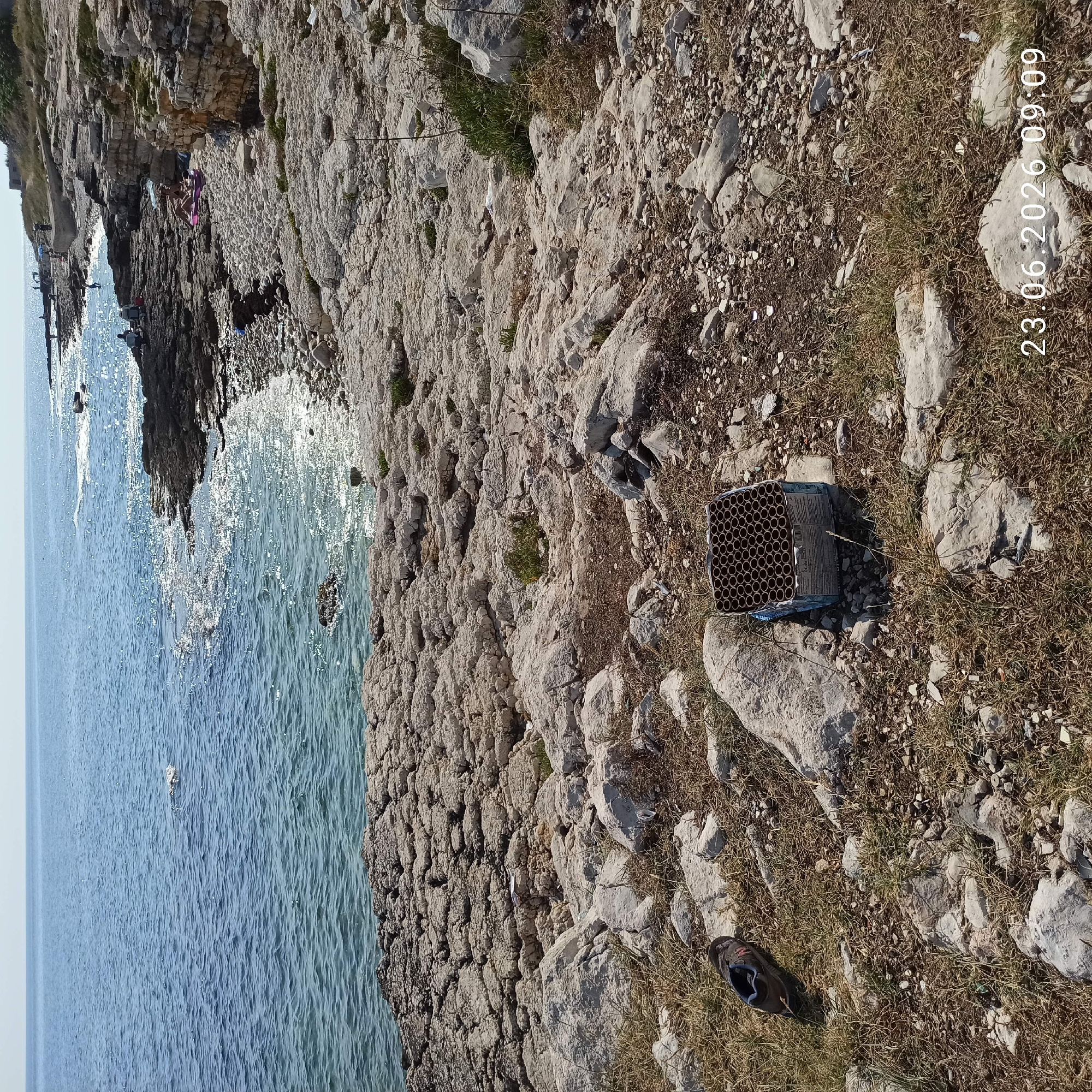 This media file depicts a rocky coastline with the sea on the left and the shore on the right. The scene is bathed in sunlight, suggesting a clear day. Several people are visible on the rocks near the water's edge, some appearing to be lounging or picnicking, indicated by colorful items that could be towels or blankets. The water is a bright blue, with sunlight reflecting off its surface.
In the foreground on the right side of the image, there is a box of fireworks sitting on the ground amidst dry grass and rocks. A single shoe is also present near the fireworks. The overall impression is of a sunny day at a coastal location, possibly a recreational spot.
Visible text in the image includes a date and time stamp in the upper right corner: "23.06.2026 09:09". The context provided suggests the location is Molfetta, Italy. The weather appears to be pleasant and warm, suitable for outdoor activities. The presence of fireworks might suggest a celebratory occasion or the aftermath of one.