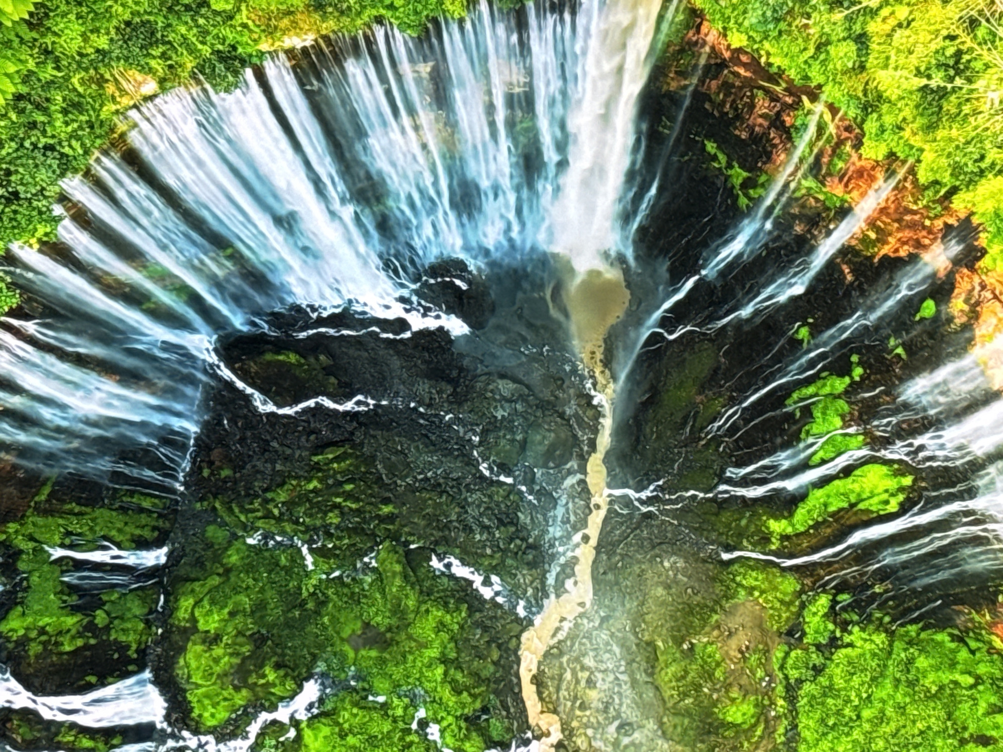 This is an aerial, high-angle, wide shot of a majestic waterfall cascading down a lush, verdant cliffside. The water, appearing bright white and translucent, fans out from the top of the frame, creating a curtain-like effect as it plunges into a dark, rocky pool below. The surrounding environment is overwhelmingly green, with dense foliage clinging to the cliff faces, suggesting a vibrant natural ecosystem. A streak of lighter colored water, possibly sediment or a different water source, snakes down the center of the waterfall's path. The image captures the raw power and beauty of nature, with the contrast between the white water, dark rocks, and vivid green vegetation being particularly striking. This scene is of the waterfall in the city of Burgas, Bulgaria.