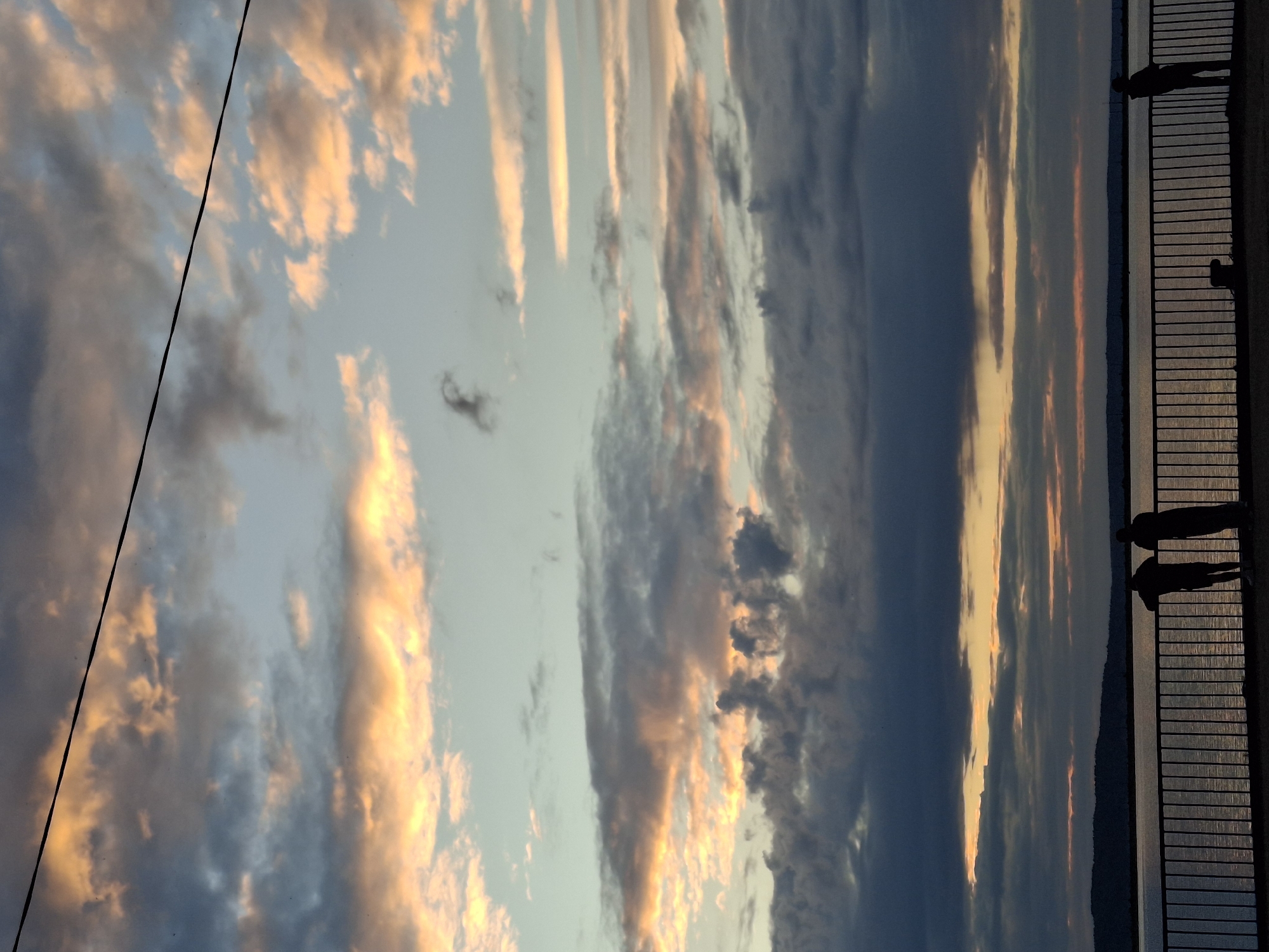 This is a landscape photograph taken at sunset over the Kama River reservoir in Chaikovsky, Russia. The sky is filled with a dramatic display of clouds, ranging from dark, heavy storm clouds to wispy, illuminated clouds tinged with orange and yellow from the setting sun. A thin black wire stretches diagonally across the upper portion of the frame. In the foreground, silhouetted figures stand by a railing, looking out at the water. The water's surface reflects the colors of the sky. A distant treeline marks the horizon. The overall mood is serene and contemplative.