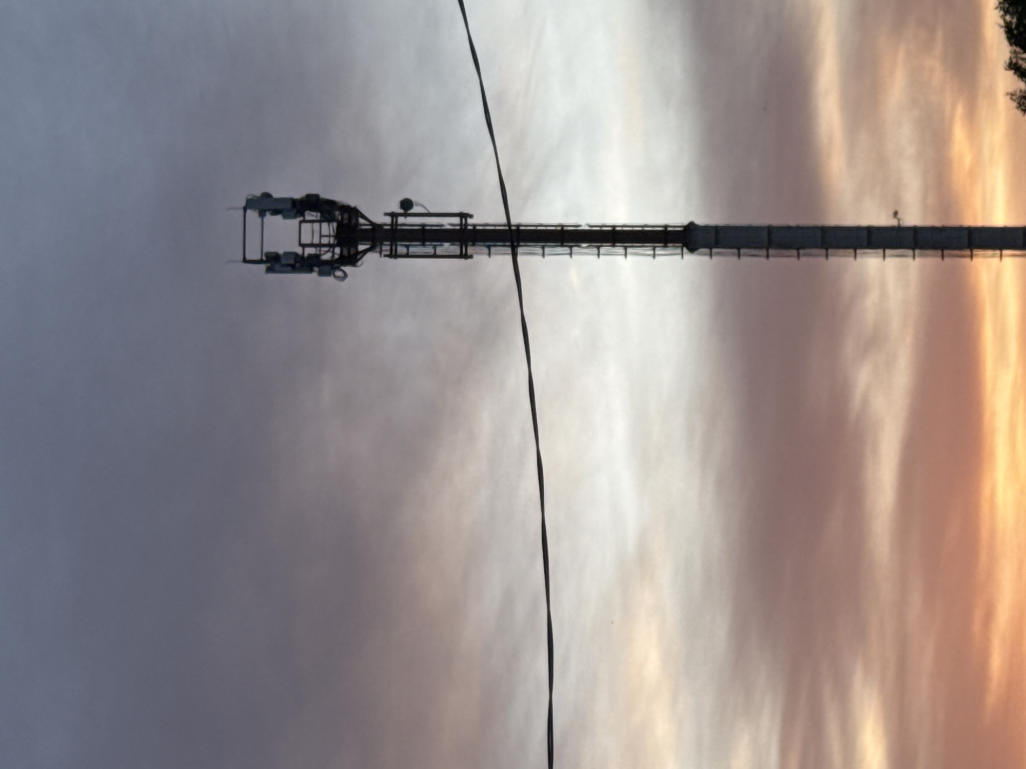 The image presents a sideways view of a communication tower against a backdrop of a sky with striated cloud patterns, transitioning from grey to orange hues. A dark, thin cable runs vertically through the frame, bisecting the tower's length. The tower appears to be constructed of a lattice-like framework with various pieces of equipment mounted at the top. The location is unknown.