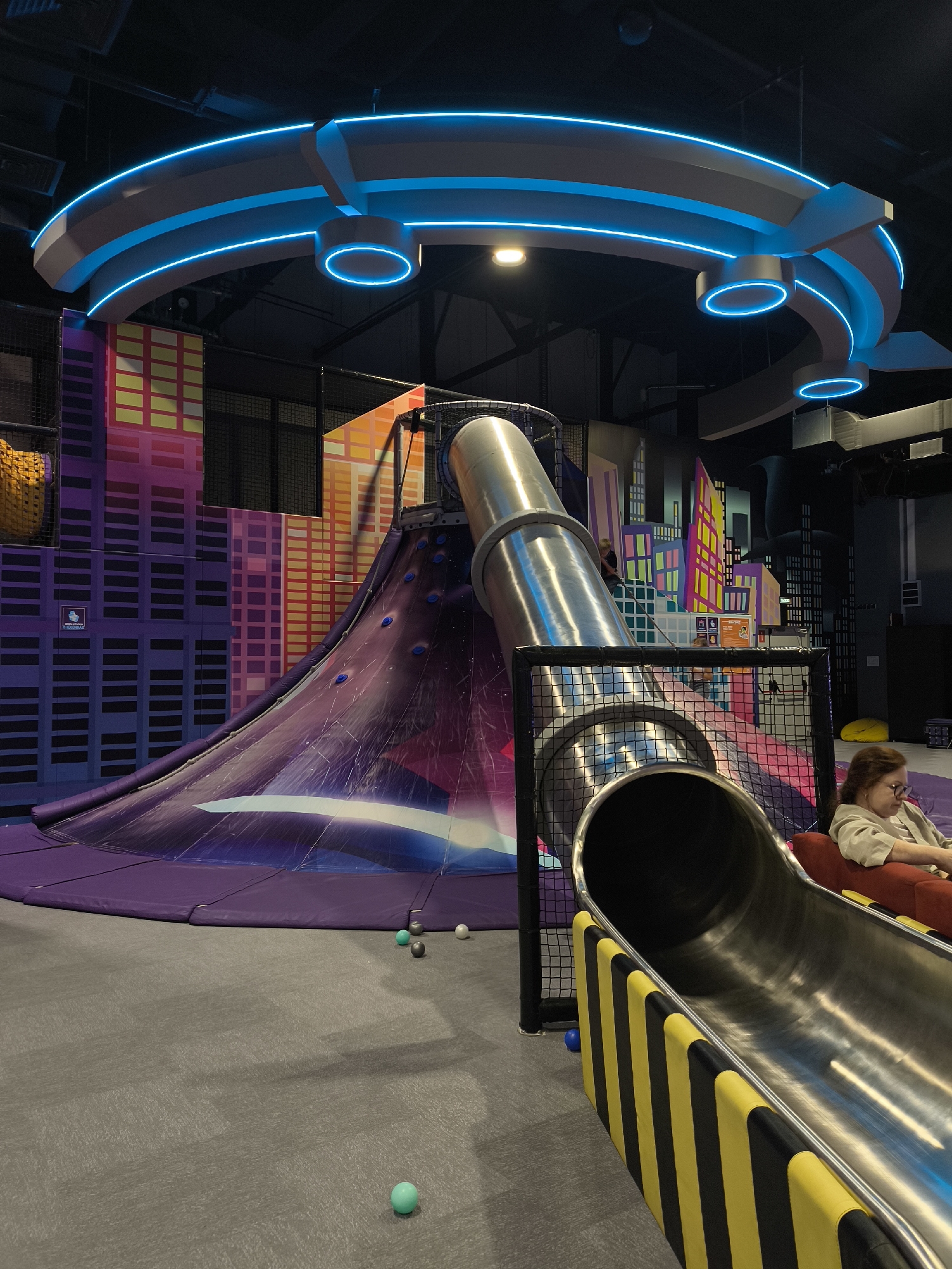 The image depicts an indoor playground with a city-themed design. A large, winding metal slide is a prominent feature, with a purple matting and a space-themed graphic at its base. The background is decorated with colorful panels resembling skyscrapers. Blue neon lights form a circular structure overhead. A woman is seated on a red couch to the right, observing the scene. Several small balls are scattered on the gray carpeted floor. There is a climbing wall integrated into the slide structure. The location is stated to be in Orlovka, Russia.
