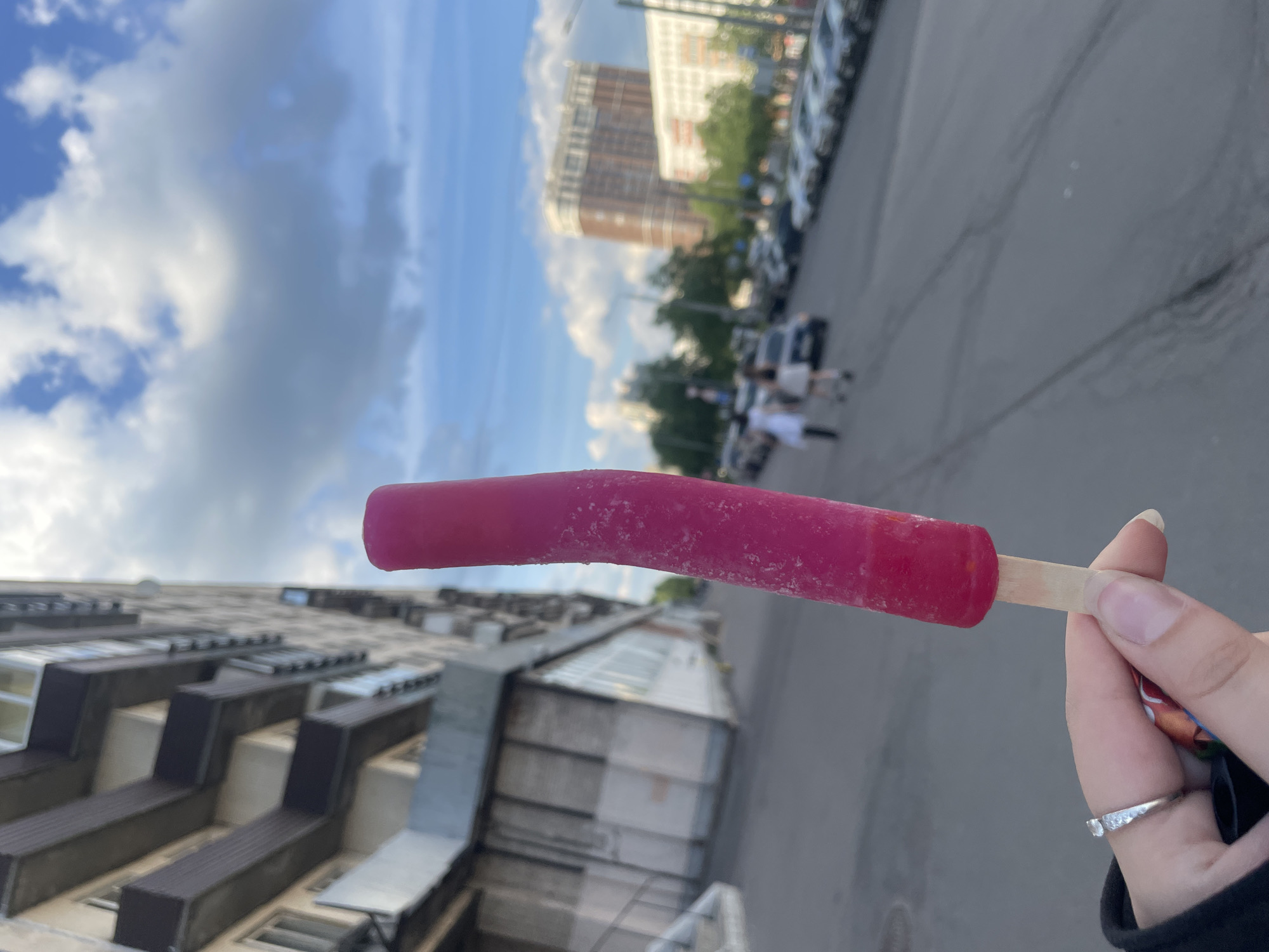 A human left hand holds a curved, fuschia-colored popsicle on a light-colored wooden stick, positioned in the lower-center to mid-right of the frame. The popsicle's surface shows small, visible ice crystals. The hand exhibits light skin tone, with a silver-colored ring worn on the ring finger.
The scene is an urban outdoor environment in Unknown, Russia. To the immediate left, a multi-story building with a light-colored concrete facade and dark-colored balconies extends vertically. The ground level of this structure features large window panels and a set of exterior concrete steps leading to an entrance.
A grey asphalt street, marked with tire traces and minor surface irregularities, runs from the lower-center towards the background. Vehicles are parked in parallel formation along the right side of this street. In the mid-ground, several individuals are visible walking on a sidewalk adjacent to the parked cars. Two figures are more distinct, one dressed in a light-colored top and dark lower garments, and another in a light-colored dress or skirt.
The distant background features multiple multi-story buildings, including a prominent brown structure and a lighter-colored building. Scattered green trees indicate the presence of vegetation along the street. The sky occupies the upper portion of the image, displaying a combination of blue sky and large, interspersed white and grey clouds. The overall lighting suggests a daytime setting.