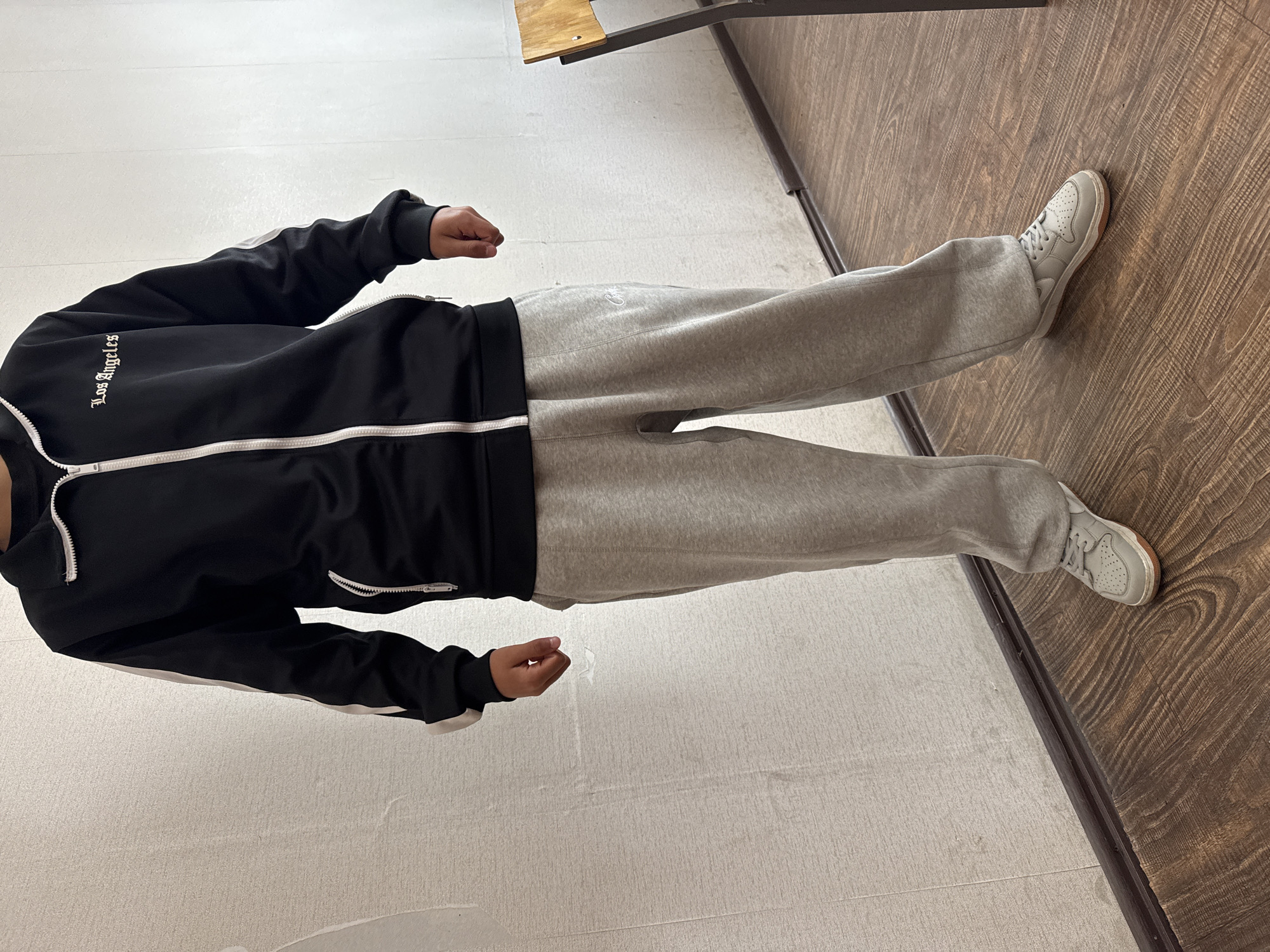 This media file captures an indoor scene in Yerevan, Armenia, focusing on a person from the neck down.
**Subjects:** The primary subject is one person, whose upper body (from the neck down), arms, and legs are visible. Their face is not shown.
**Objects:** The person is wearing a black tracksuit jacket with white stripes along the sleeves and a white zipper, grey sweatpants, and light grey sneakers with white laces. The background includes a wooden floor and a lighter, possibly carpeted or tiled, floor, with a clear transition line between the two. Part of a metal chair or desk frame with a wooden seat/surface is visible in the upper right corner.
**Scene/Setting:** The setting is indoors, characterized by artificial lighting and two distinct flooring types. The presence of a desk or chair suggests a classroom, office, or similar institutional environment.
**Activity or event taking place:** The person appears to be standing still, possibly posing for the photograph or waiting. Their arms are slightly bent, and their hands are clenched into loose fists.
**Notable details:**
* **Time of day/weather:** Not discernible from the indoor environment.
* **Location cues:** The flooring and furniture suggest an interior space.
* **Emotions:** Cannot be determined as the subject's face is not visible.
* **Attire:** The clothing is casual and athletic, indicating comfort.
**Visible text:** The left sleeve of the black jacket features white text that reads "Los Angeles" in an Old English or Gothic script. A small, indistinct white logo or text is visible on the upper left thigh of the grey sweatpants.