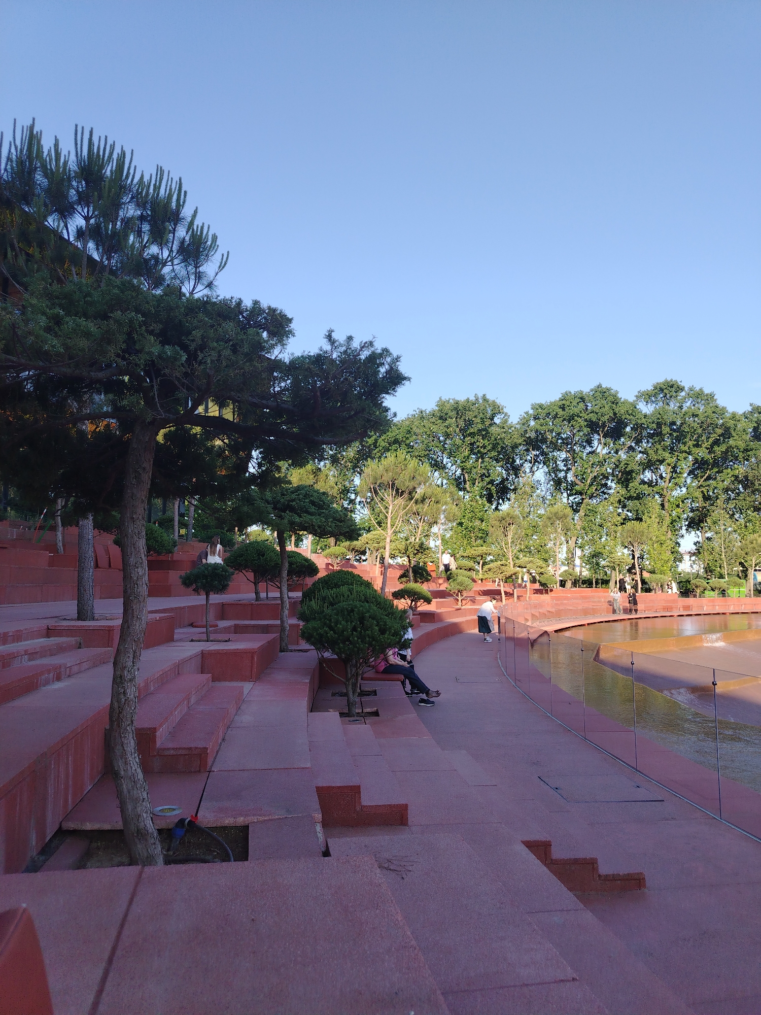 The image depicts an outdoor amphitheater with tiered red stone seating. A variety of trees, including pine and deciduous varieties, are planted throughout the seating area and in the background. A shallow water feature with a light brown surface is visible to the right, bordered by a glass railing. Several individuals are present in the scene. In the foreground, a person sits on a bench. Further back, a person in white walks, and a few others are seen standing near the water feature. The sky is clear and blue, indicating daylight. The architecture suggests a modern park or public space designed for performances or gatherings. While not explicitly identifiable from the image alone, this type of landscaped public space is consistent with features found in cities like Krasnodar, Russia, known for its parks and recreational areas.