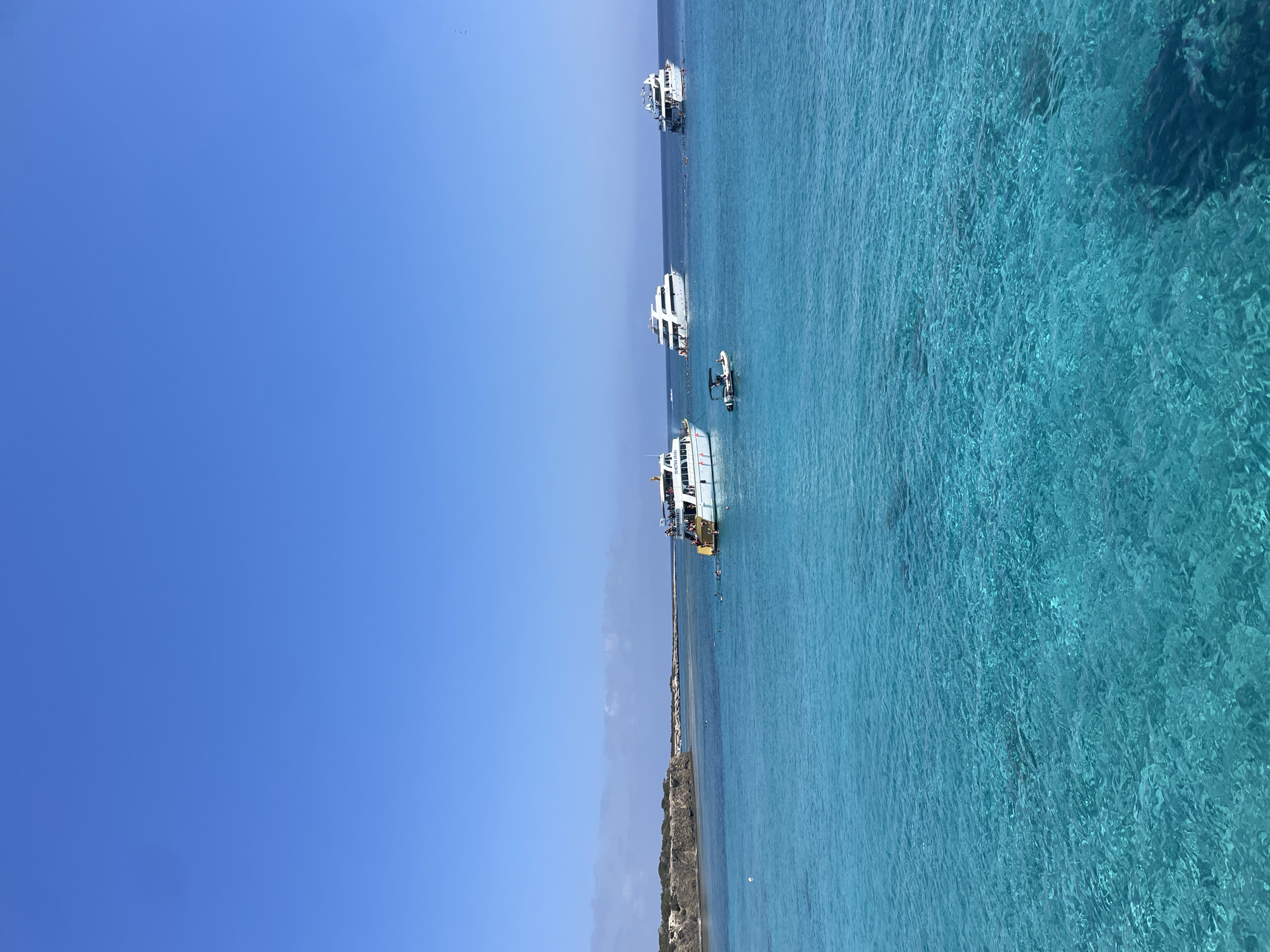 The image depicts a clear, bright day at sea near Neo Chorio, Cyprus. The foreground is dominated by exceptionally clear, turquoise water, allowing the rocky seabed to be visible. In the midground, several white boats of varying sizes are anchored or cruising. One larger white boat on the right side of the frame has people on its deck and in the water around it. Another large white boat is positioned further to the left, with a smaller dinghy in front of it. A third, smaller white boat is visible to the far left. In the background, a rocky coastline with sparse vegetation stretches across the horizon. The sky is a vast expanse of clear, light blue. The overall impression is one of a serene and inviting coastal environment.