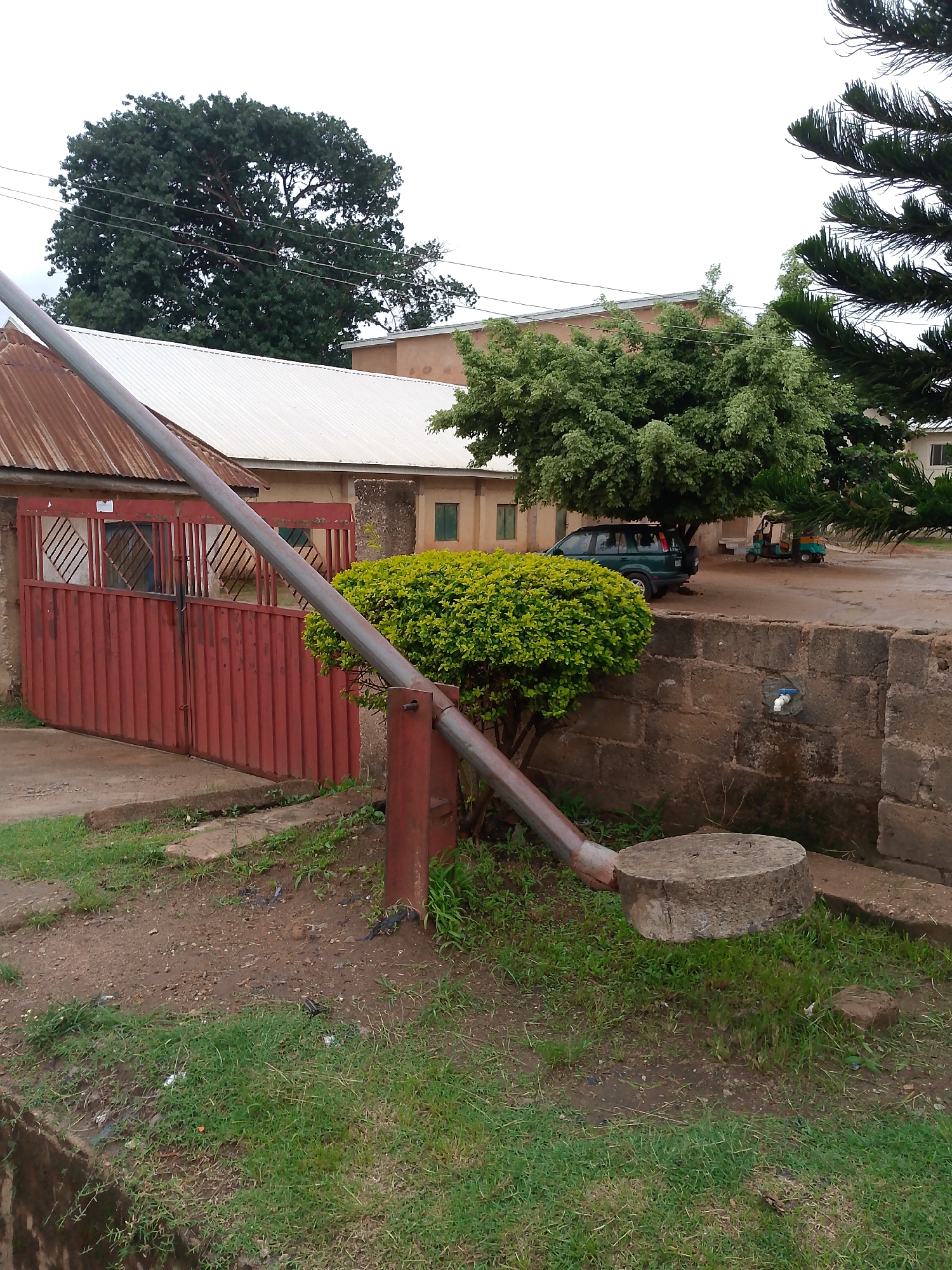The image presents an outdoor scene depicting an entrance or boundary within a property, likely in Bukuru, Nigeria. In the foreground, a dark red metal barrier arm extends diagonally from a sturdy post on the left to a cylindrical concrete block resting on the ground to the right, suggesting controlled access. The ground beneath the barrier is a mix of dirt and patches of green grass.
Behind the barrier, to the left, a red metal gate with a decorative diamond lattice pattern secures an entry point to a compound. Adjacent to the barrier, a dense, round green bush thrives. To the right of the bush, a low, unplastered concrete block wall features a white plastic faucet with a blue handle.
In the midground, several buildings are visible. The one directly behind the gate has a rusty, corrugated metal roof. A larger building further back has a newer, silver-colored corrugated roof. Under the canopy of a large, leafy green tree, a dark green SUV is parked, and further in the background, a three-wheeled auto-rickshaw (locally known as a keke Napep) is also visible. Another tall, evergreen tree with needle-like leaves frames the right side of the image.
The scene appears to be set during daytime under an overcast or cloudy sky, indicated by the diffused lighting and lack of strong shadows. No people are visible, and there is no discernible text on any of the objects or structures. The overall impression is one of a functional, semi-developed compound entrance.