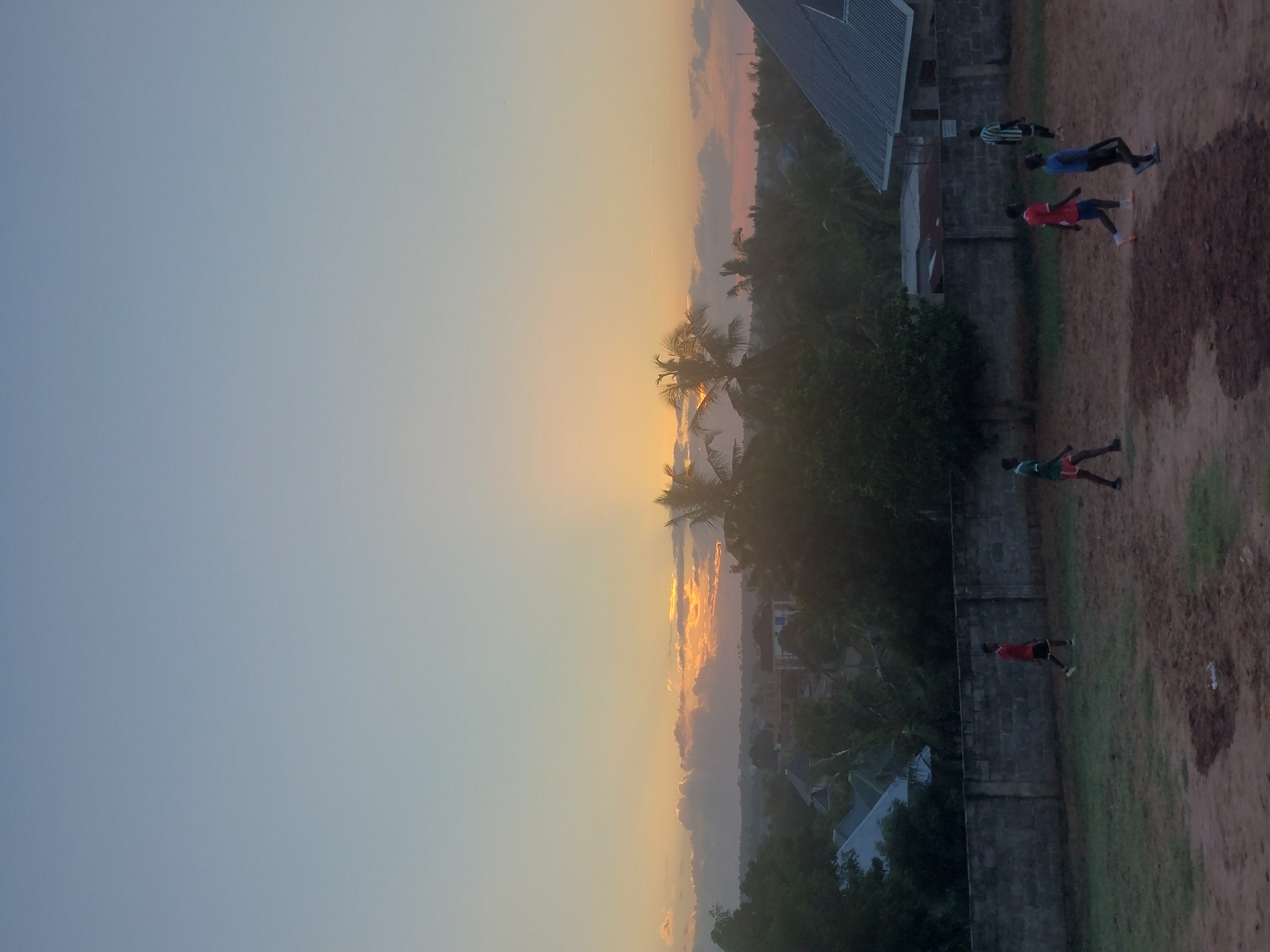 The image captures a scene in Dar es Salaam, Tanzania, at dawn or dusk, as indicated by the soft, diffused sunlight. The sky transitions from a pale blue to a warm orange/yellow, suggesting either sunrise or sunset. The foreground shows a partially grassy, partially dirt field where at least four individuals are present. They appear to be running or exercising. In the background, palm trees and other vegetation are visible. Buildings are interspersed within the trees, and a wall divides the field from the developed area. The scene is illuminated by the sun on the horizon.