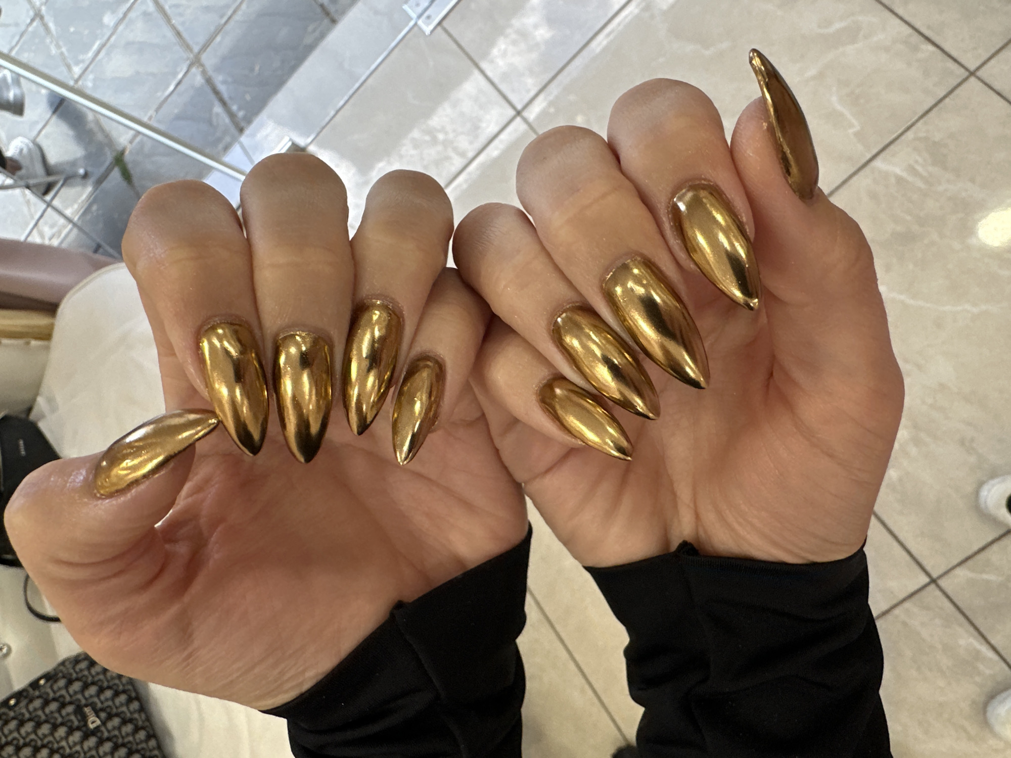 The image shows two hands with gold stiletto nails held up against a background of light-colored floor tiles. The person is wearing a black long-sleeved garment. There is a glimpse of a white cushioned chair or bench and part of a black patterned Dior handbag on the left. A leg with a white sneaker is seen on the upper left corner. The location appears to be an indoor space. The city of Sofia, Bulgaria is not evident from the image.