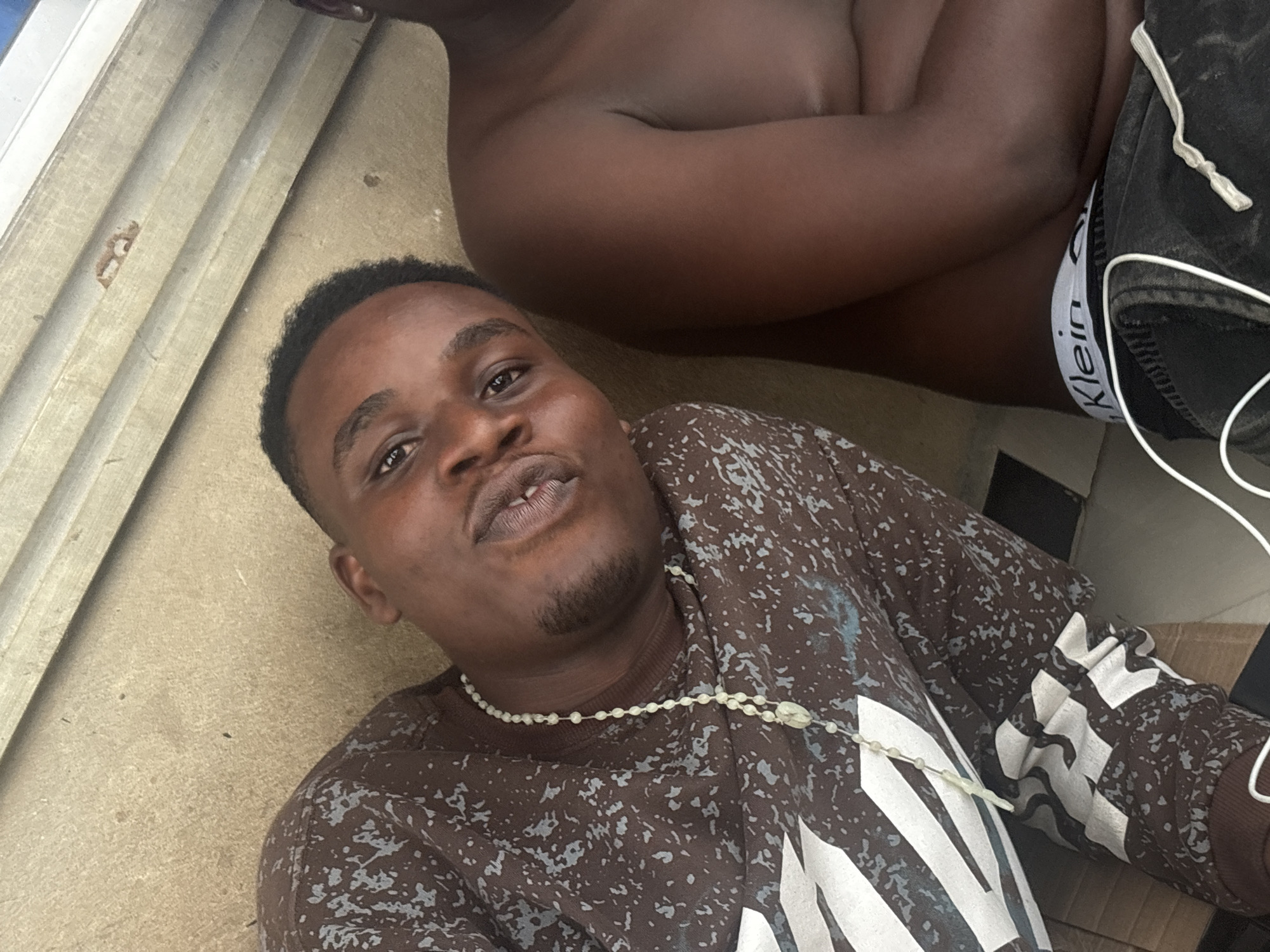 A medium shot depicts a young, dark-skinned man with short, dark hair and a close-trimmed beard. He is wearing a brown and white patterned long-sleeved shirt and a white beaded necklace. He is looking directly at the camera with a slight smirk. Behind him is another person, shirtless, with dark skin wearing black shorts with a visible white Calvin Klein waistband. The background includes a beige wall and glimpses of an interior setting. The location, Ikot Ekpene, Nigeria, is not discernible from the image itself.