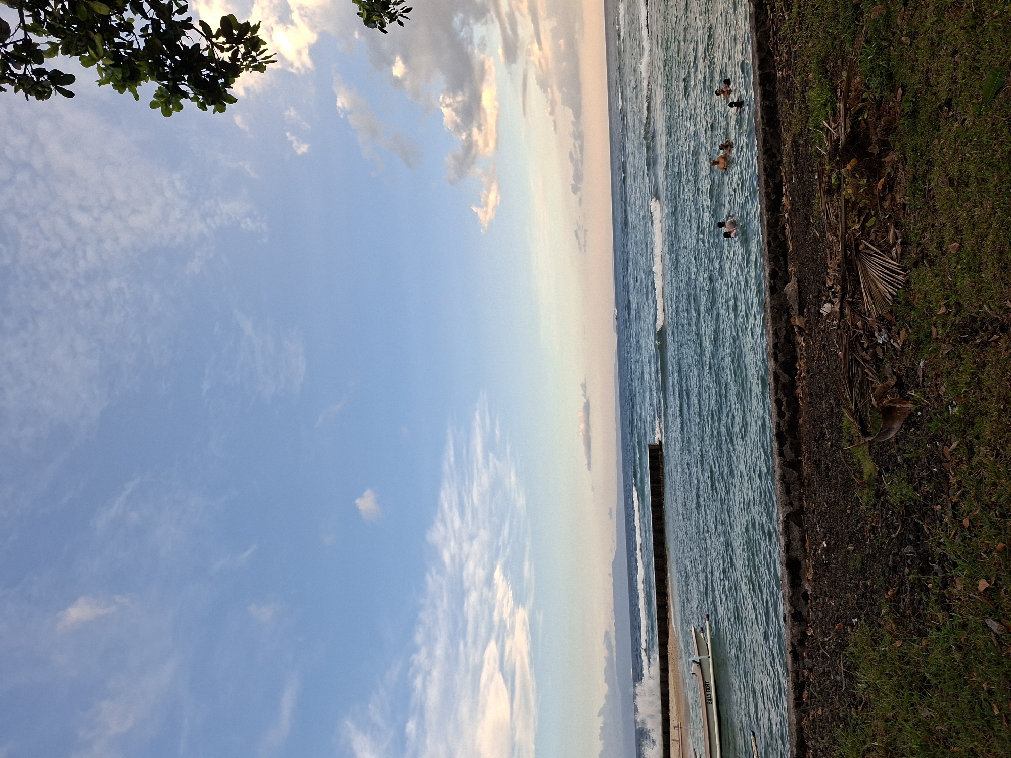 The image shows a scene at the beach, likely in Karangasem, Indonesia, as evidenced by the location and environment. It is oriented sideways, with the sky and scattered clouds occupying the left side of the frame and the ocean on the right. The sky transitions in color from blue to hints of pink and orange near the horizon.
In the ocean, several people are visible, appearing to be swimming or standing in the water near the shoreline. The water shows signs of waves and movement.
A strip of land or beach is visible between the ocean and what appears to be a grassy area or edge of a park, providing a thin separation between the sand and the vegetation. The foliage includes grass and scattered dead leaves or debris.
A structure, possibly a pier or part of a boat, is located near the edge of the water. The color of the ocean is light blue and green.
A tree's branches and leaves appear in the upper left corner of the image, adding a natural element to the composition.
